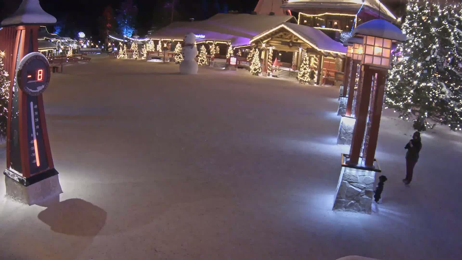 A snow-covered village square at night, illuminated by Christmas lights, features a snowman, several decorative light posts, and a person with a small dog standing near a tree.