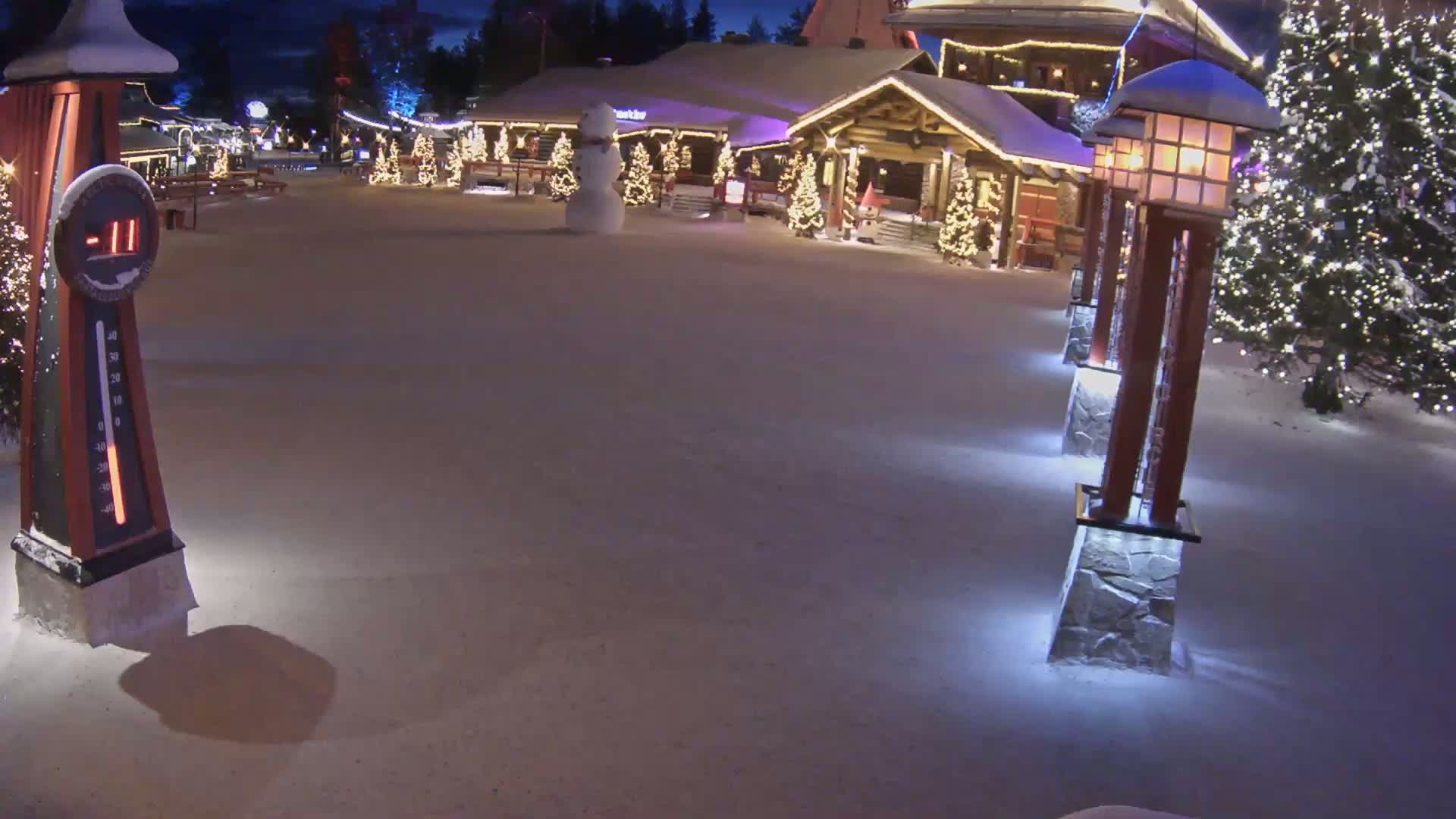 A snow-covered village square at night, illuminated by lights on buildings and lampposts, shows a snowman and Christmas trees.