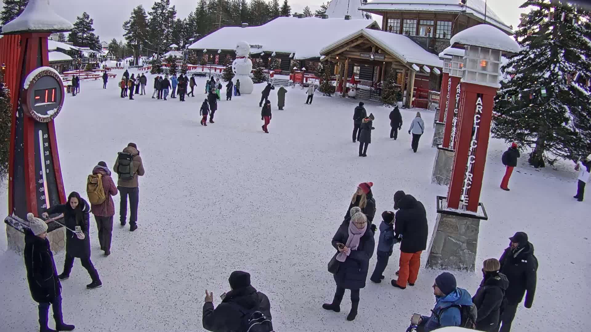 A snowy plaza with many people, a large snowman, and several tall red posts, with a building in the background under an overcast sky.