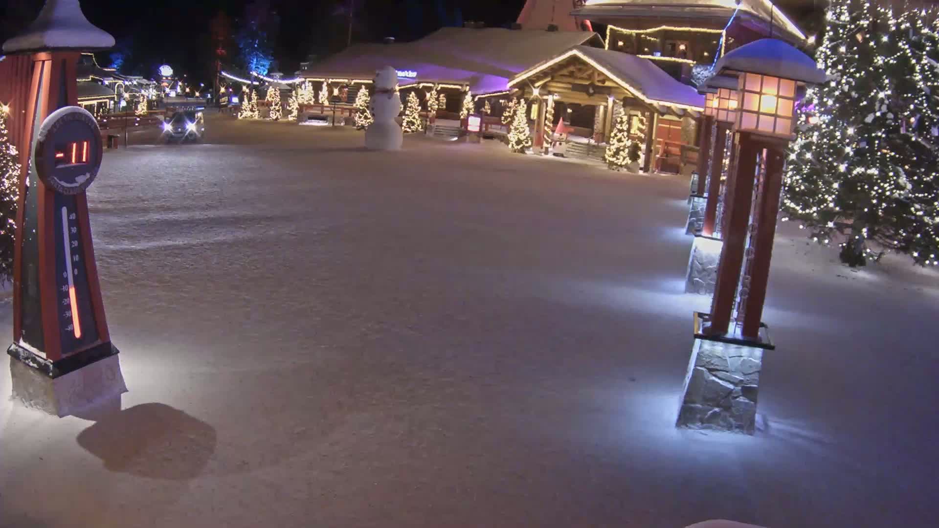 A snow-covered village square at night, illuminated by lights on buildings and lampposts, features a snowman and shows a temperature of -11 degrees.