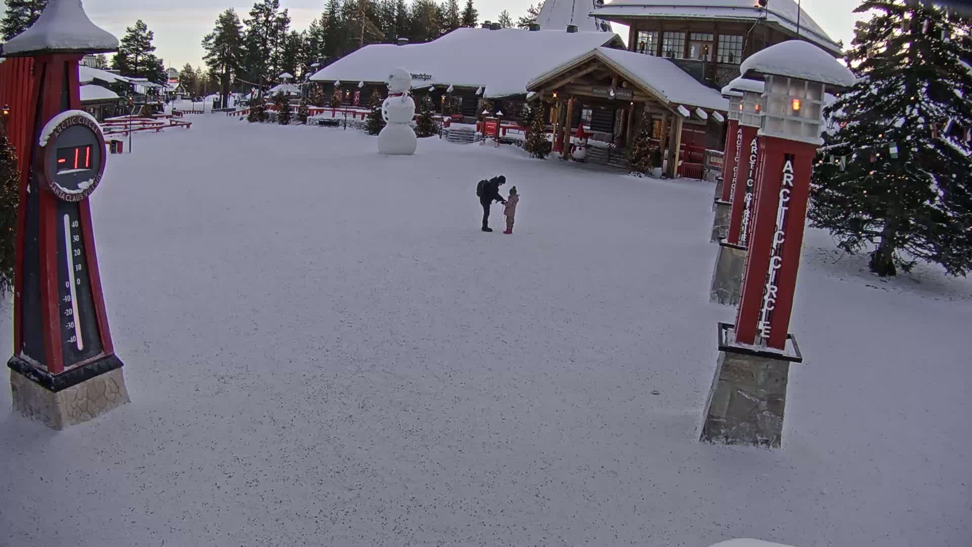 A snow-covered plaza with a building in the background, a snowman, and two people standing together; the temperature is -11 degrees.