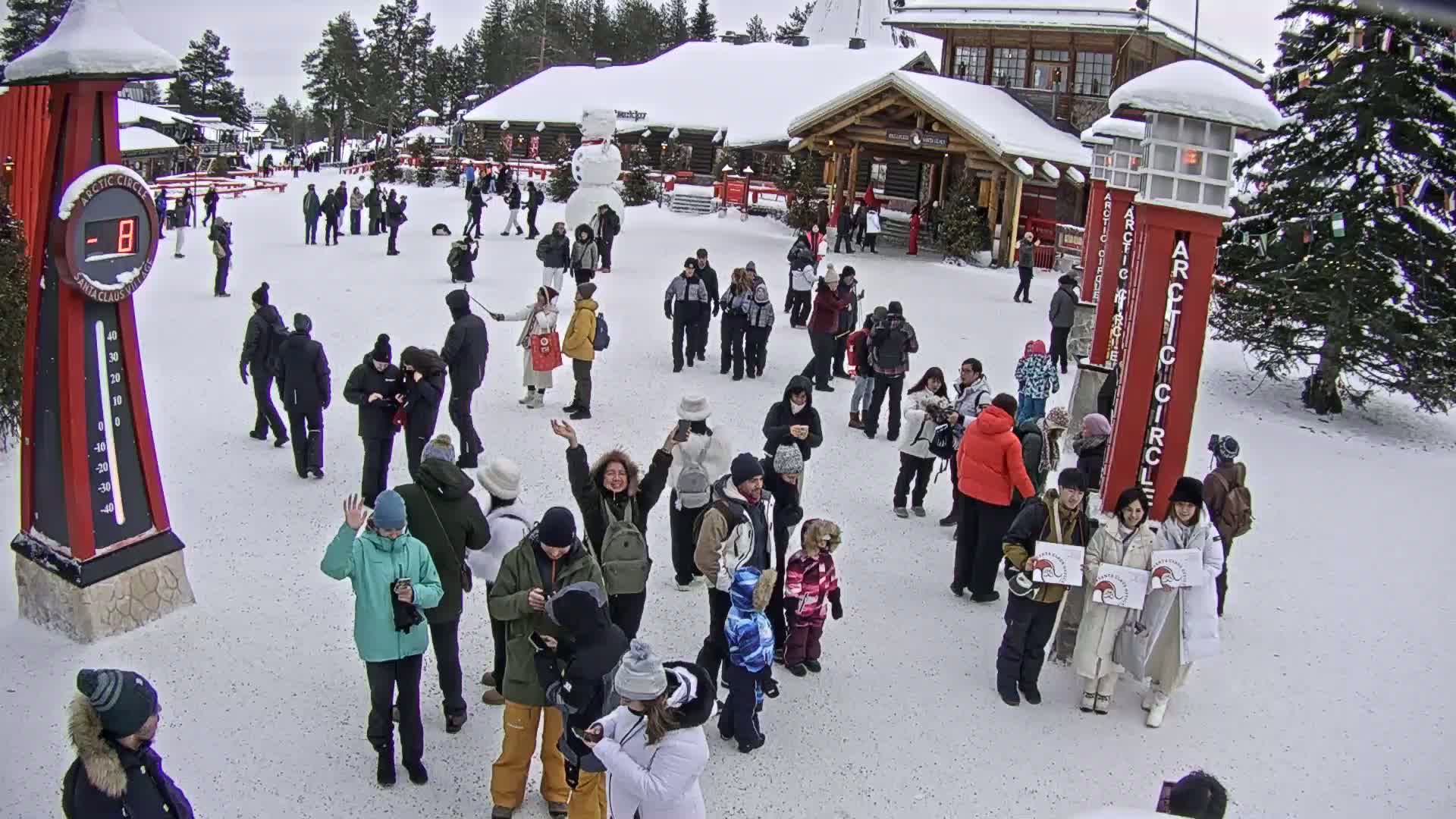 A crowd of people gathers in a snowy plaza in front of wooden buildings, a snowman, and tall red Arctic Circle signs, with a thermometer displaying a temperature below zero.