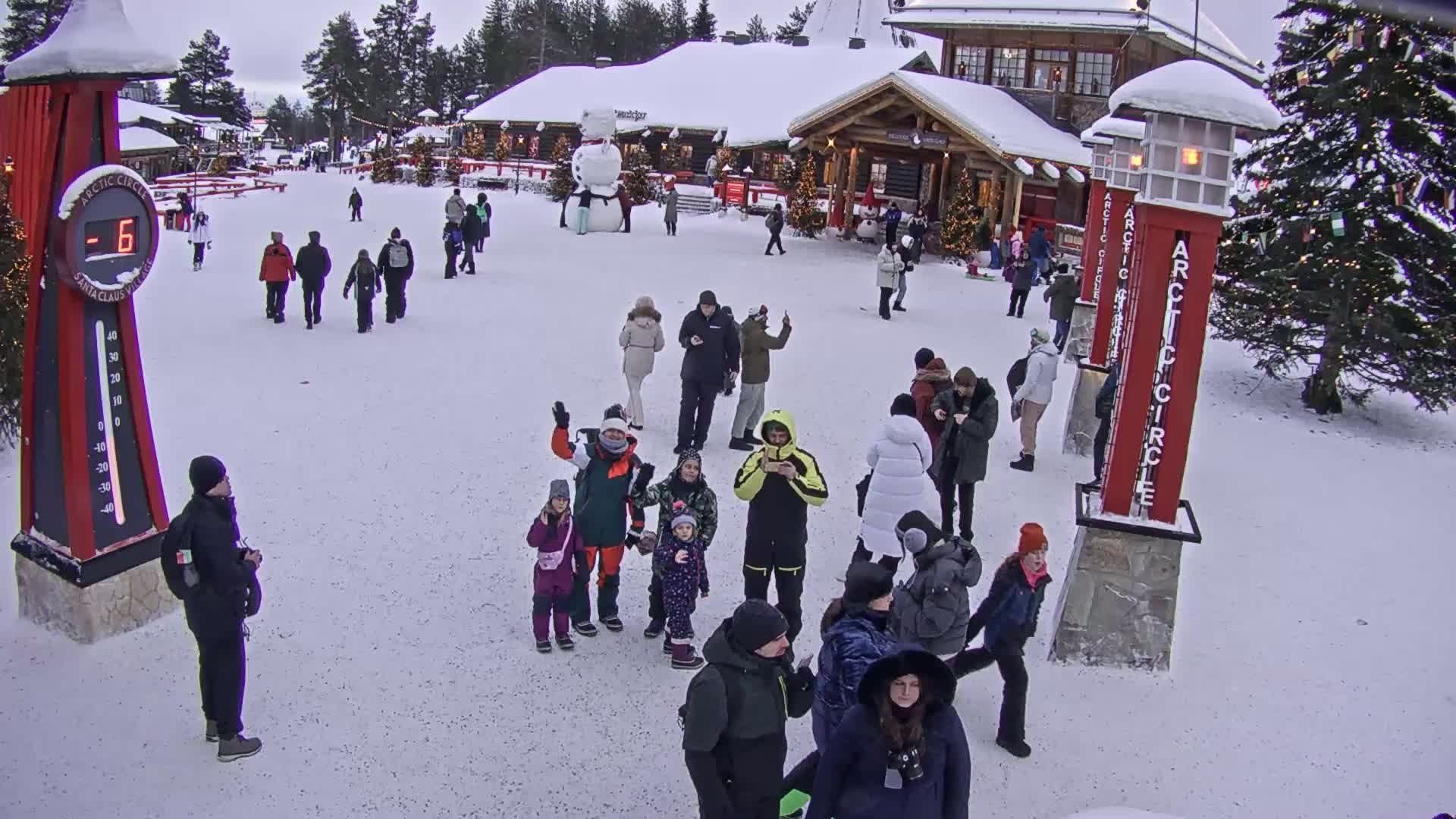 A snowy plaza in front of a building, filled with people walking around, some standing in small groups, with a large snowman visible in the background and a temperature display showing -6 degrees.