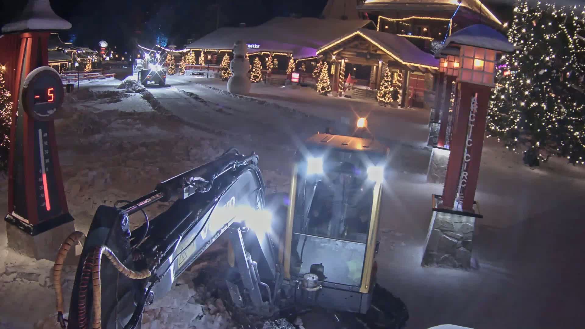 A small excavator is working in a snowy area at night near a building decorated for the holidays, with the temperature shown as -5 degrees.