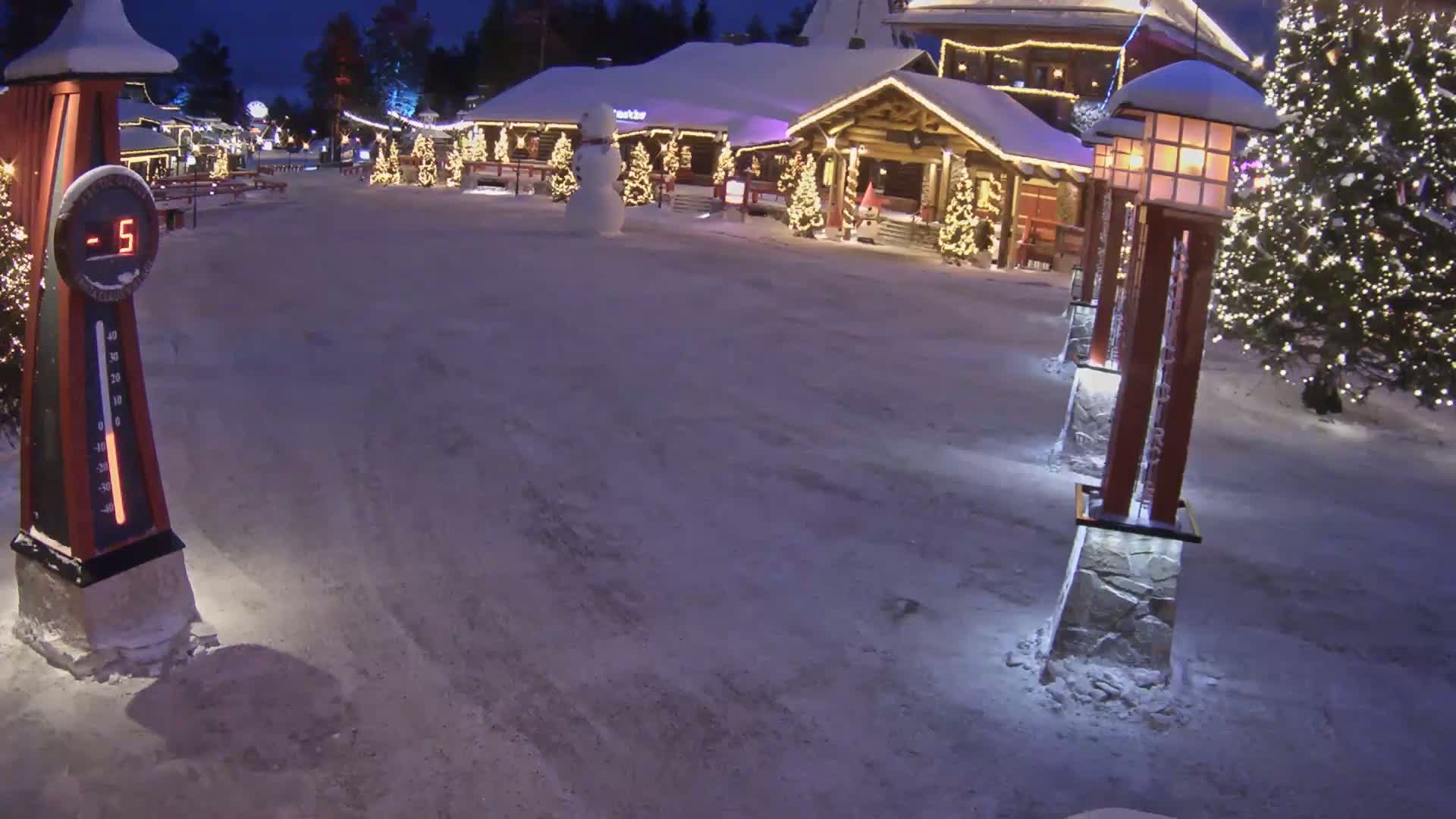 A snow-covered village square at night, illuminated by lights on buildings and lampposts, features a snowman and shows a temperature of -5 degrees.