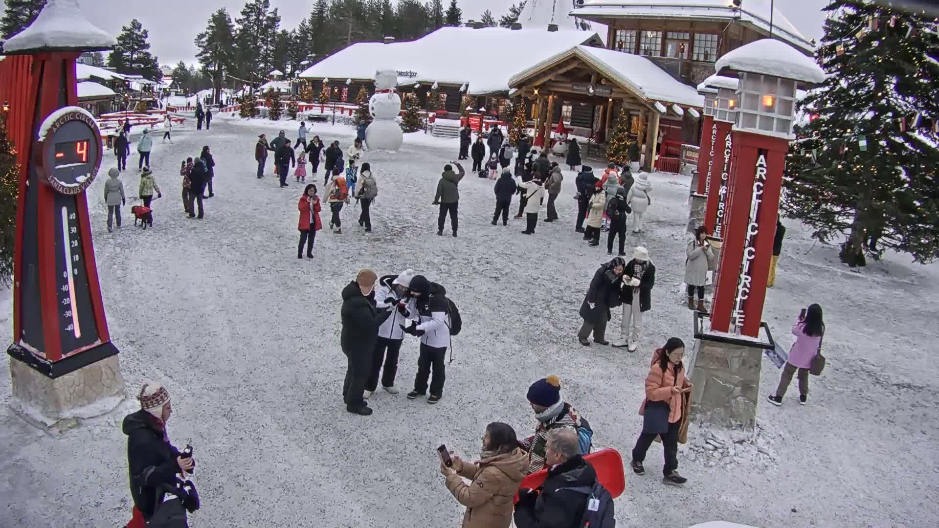 Many people are gathered in a snowy plaza in front of a building, with a large snowman nearby and a temperature of -4 degrees displayed on a nearby sign.
