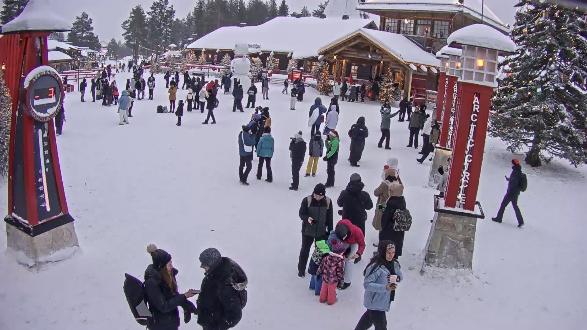 A snowy village square with many people milling about near wooden buildings, a snowman, and tall red posts.