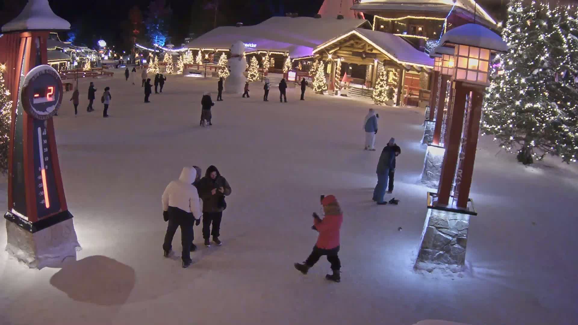 A snow-covered plaza at night, illuminated by lights, features several people walking and standing around a large snowman near a building, with a thermometer showing a temperature of -2 degrees.