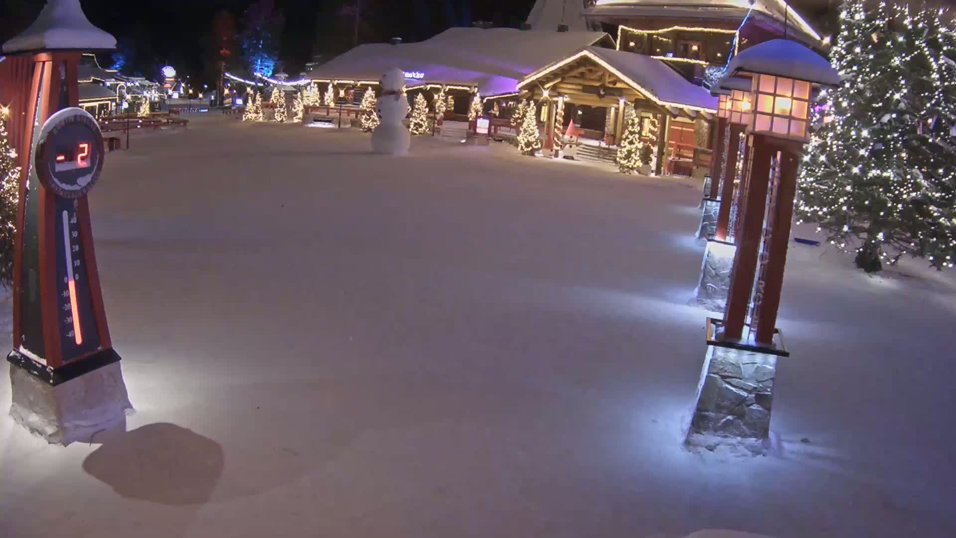A snow-covered plaza at night features a snowman, illuminated Christmas trees, and festively decorated buildings, with a thermometer indicating a temperature of -2 degrees.