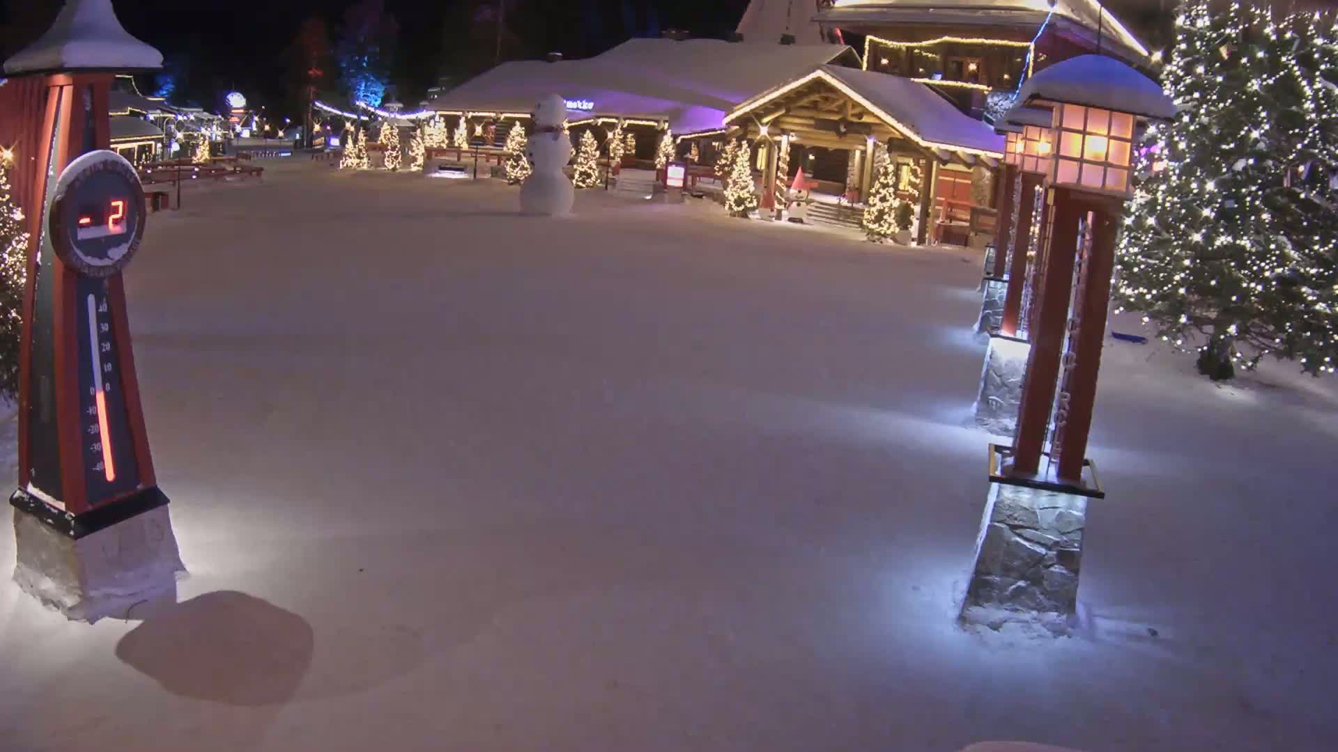 A snow-covered plaza at night, featuring a snowman, illuminated Christmas trees, and buildings with warm lighting, shows a temperature of -2 degrees on a nearby thermometer.