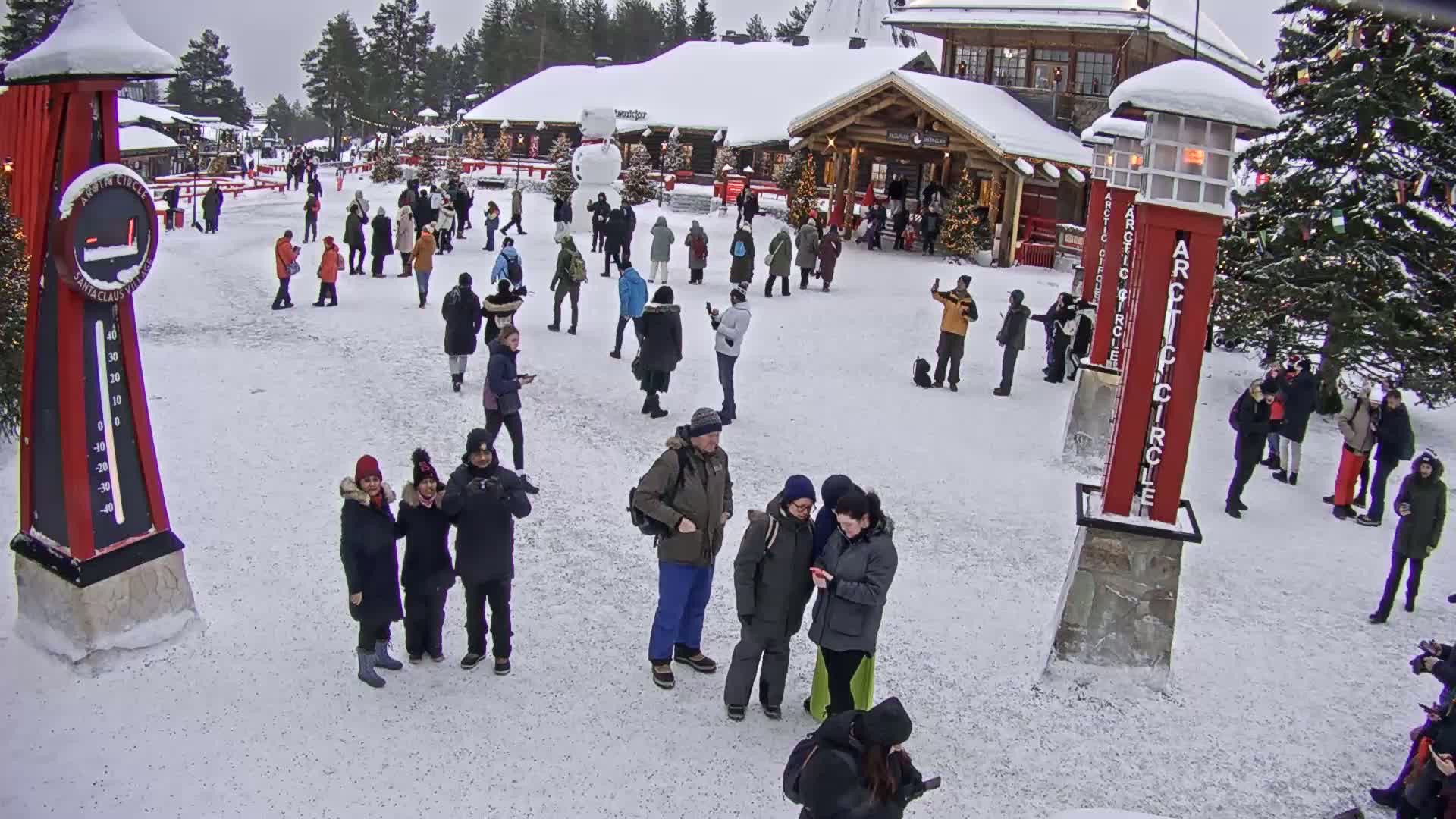 A snowy outdoor plaza features a crowd of people gathered near wooden buildings, with several tall red markers standing in the snow.