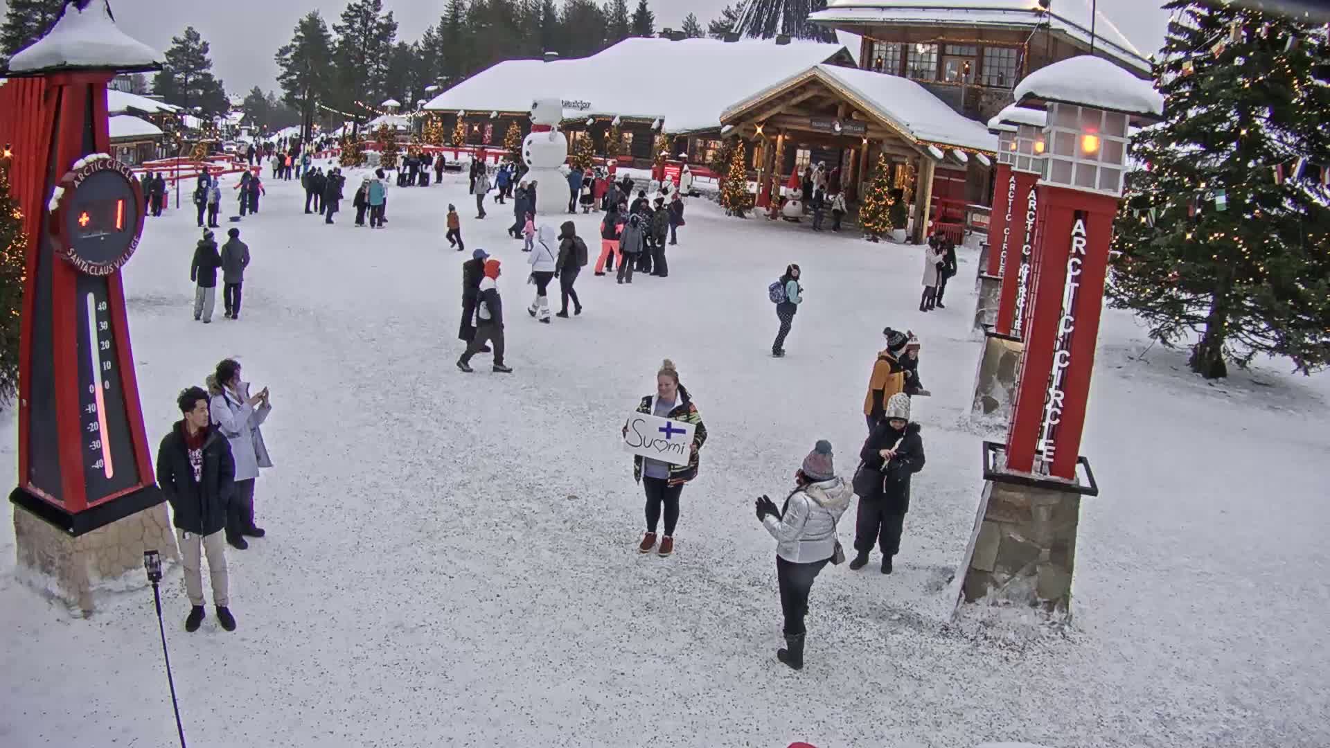 A snow-covered village square features numerous people walking around buildings decorated for the holidays under an overcast sky.