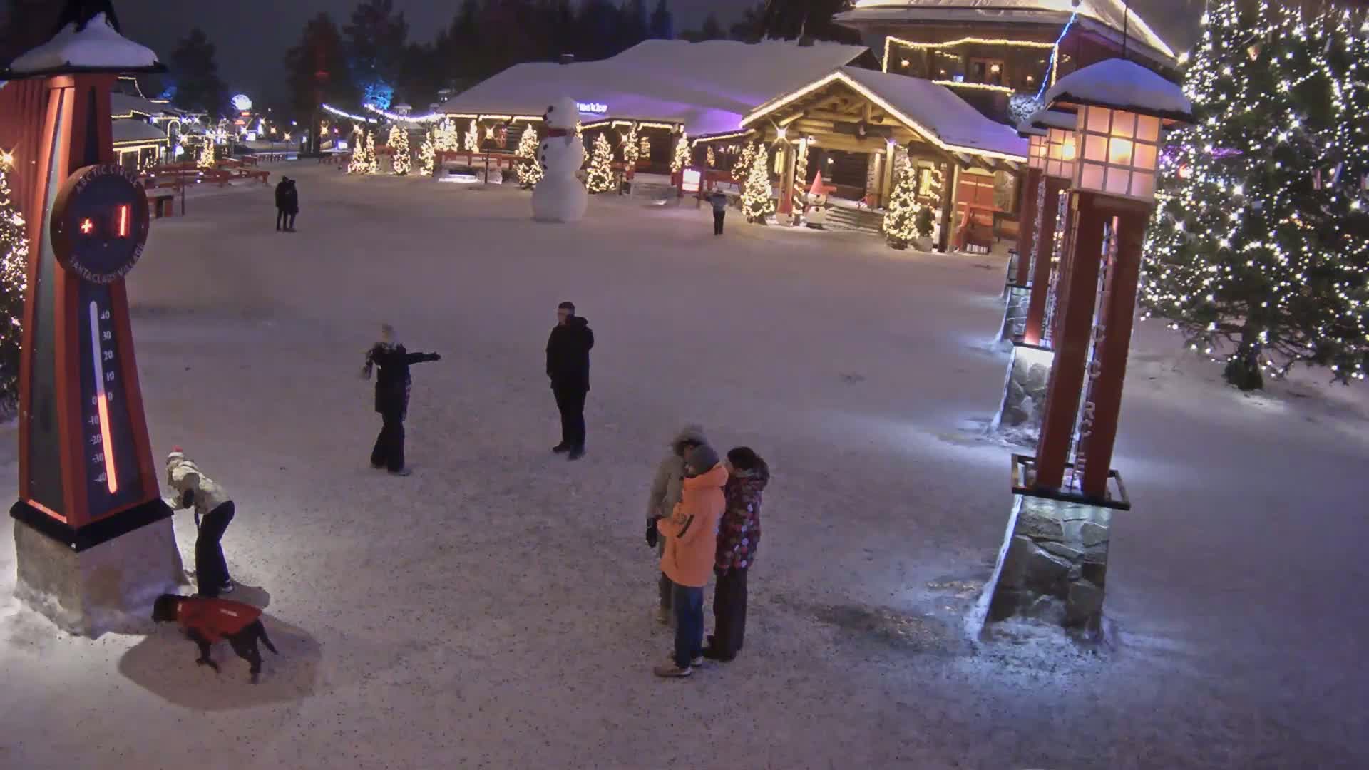 A snow-covered village square at night, illuminated by Christmas lights, features several people, a snowman, and a large thermometer.