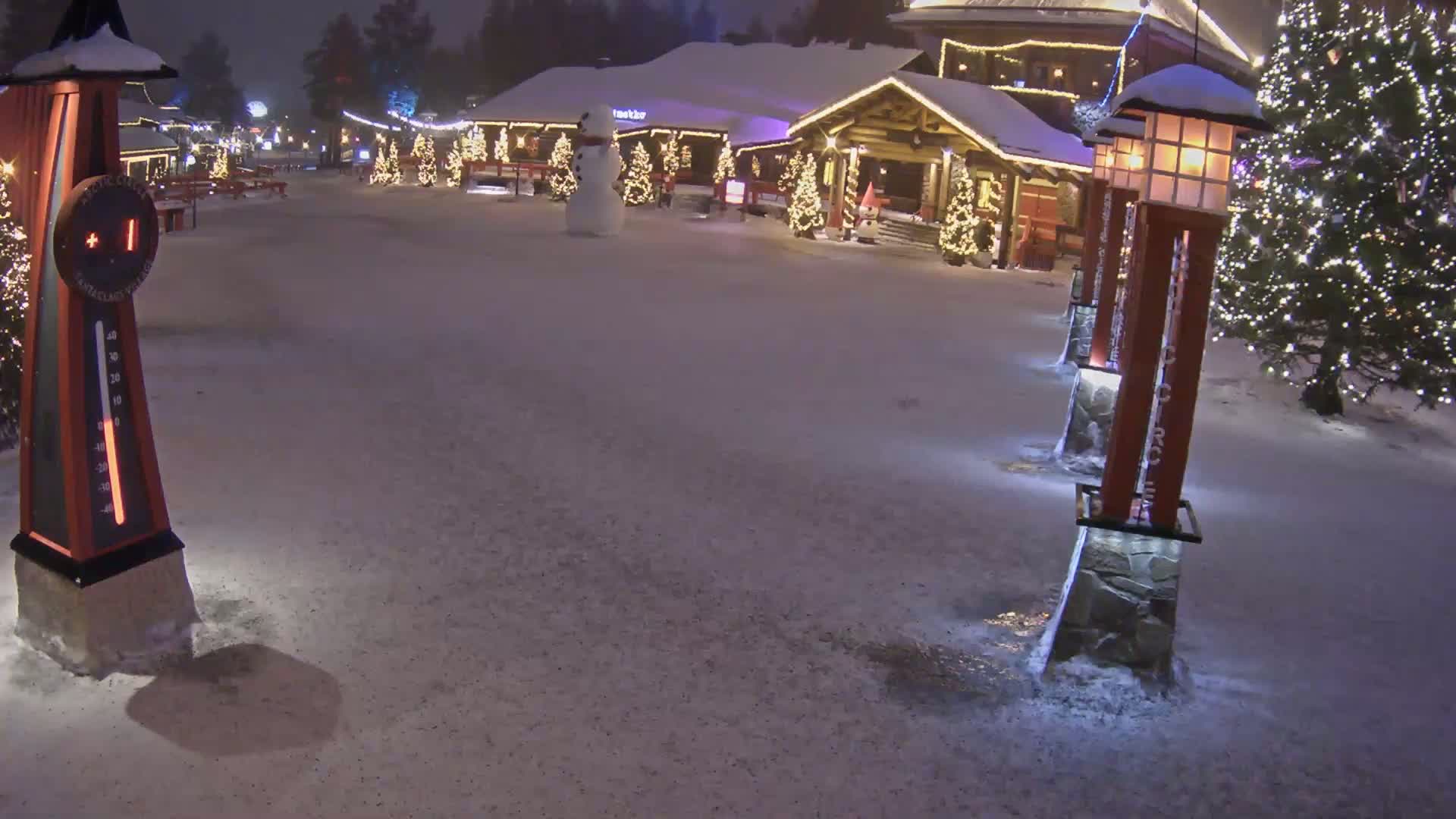 A snow-covered plaza at night features a wooden building with Christmas lights, a snowman, and several illuminated lamp posts.
