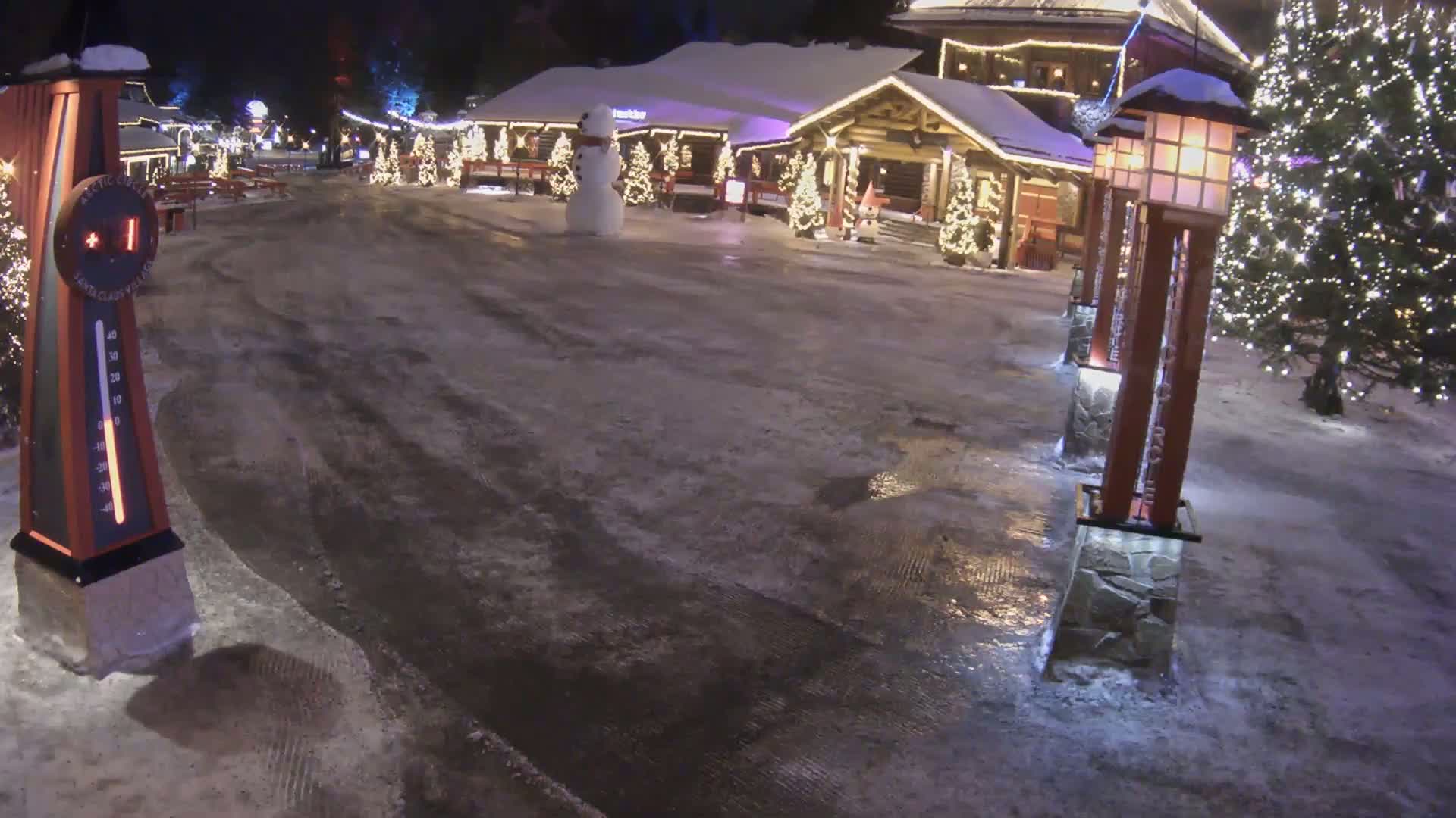 A snow-covered village square at night features a snowman, illuminated Christmas trees, and decorative lights, with patches of melted snow and ice on the ground.