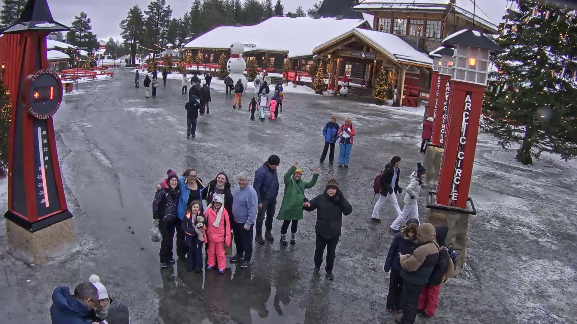 A group of people stand in a snowy plaza near buildings decorated for Christmas, under an overcast sky.
