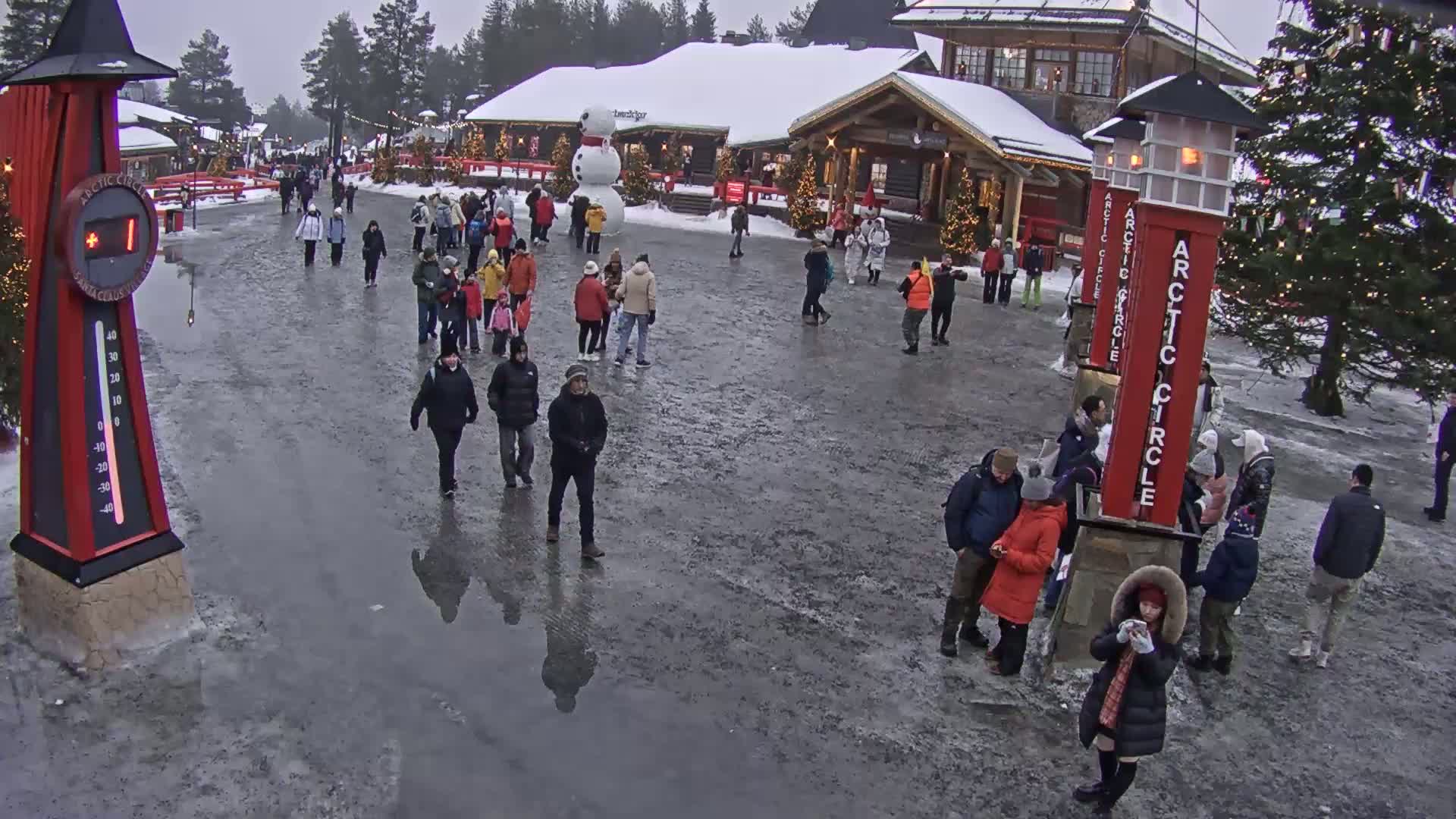 A crowd of people walk across a slushy, icy plaza in front of snow-covered buildings on an overcast day.