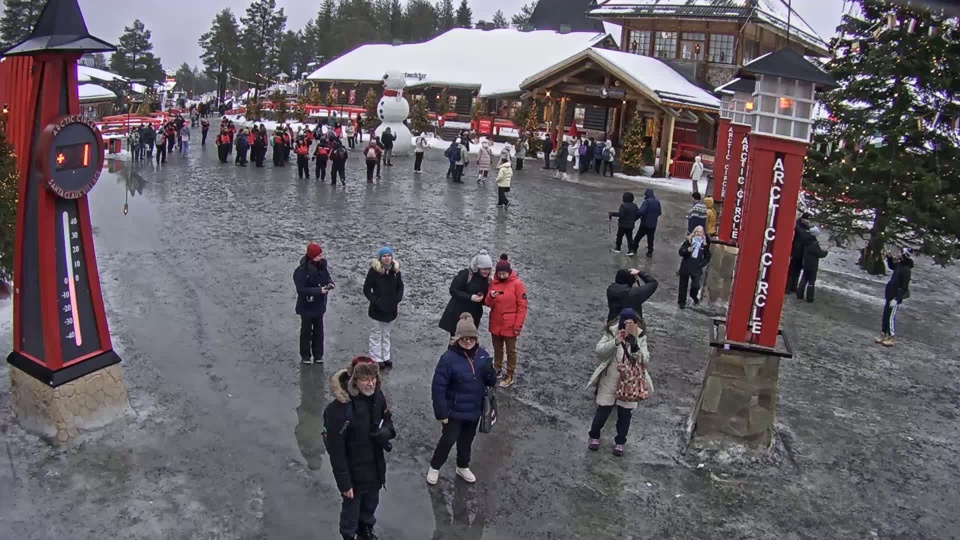 A mostly icy, open area with numerous people standing around in winter weather, near some buildings and Christmas trees.