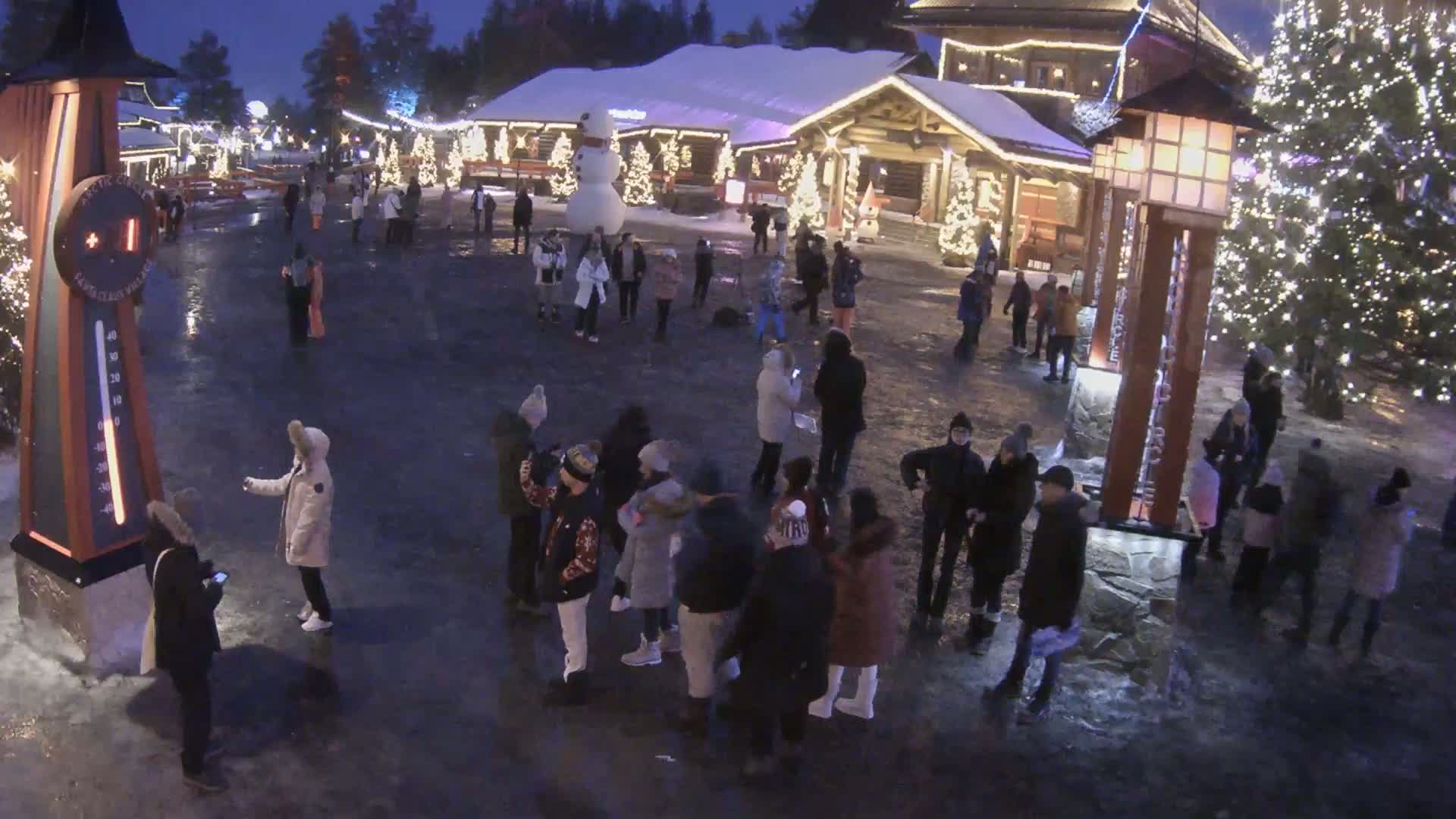 A nighttime, outdoor scene shows a crowd of people gathered in a snow-covered plaza, near a large snowman and illuminated Christmas trees.