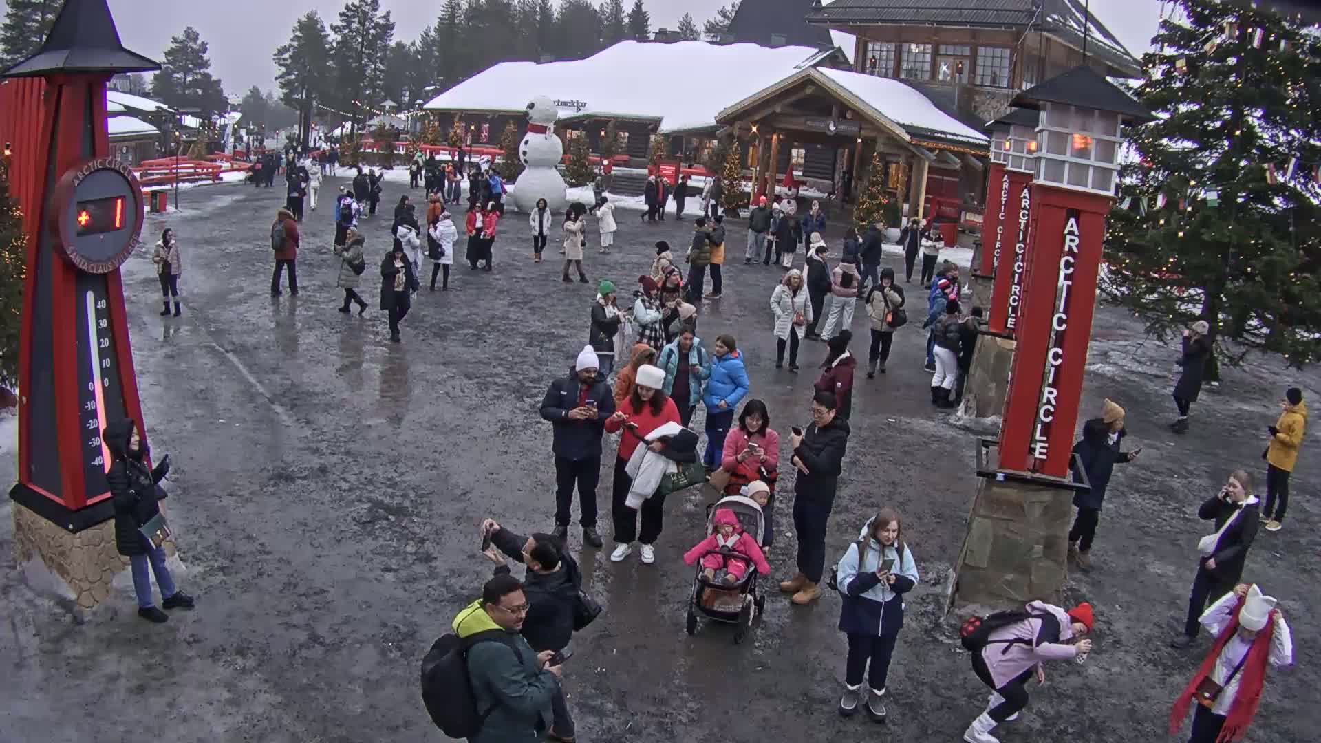 A large group of people gathers in a snowy outdoor plaza near wooden buildings, some using their phones.