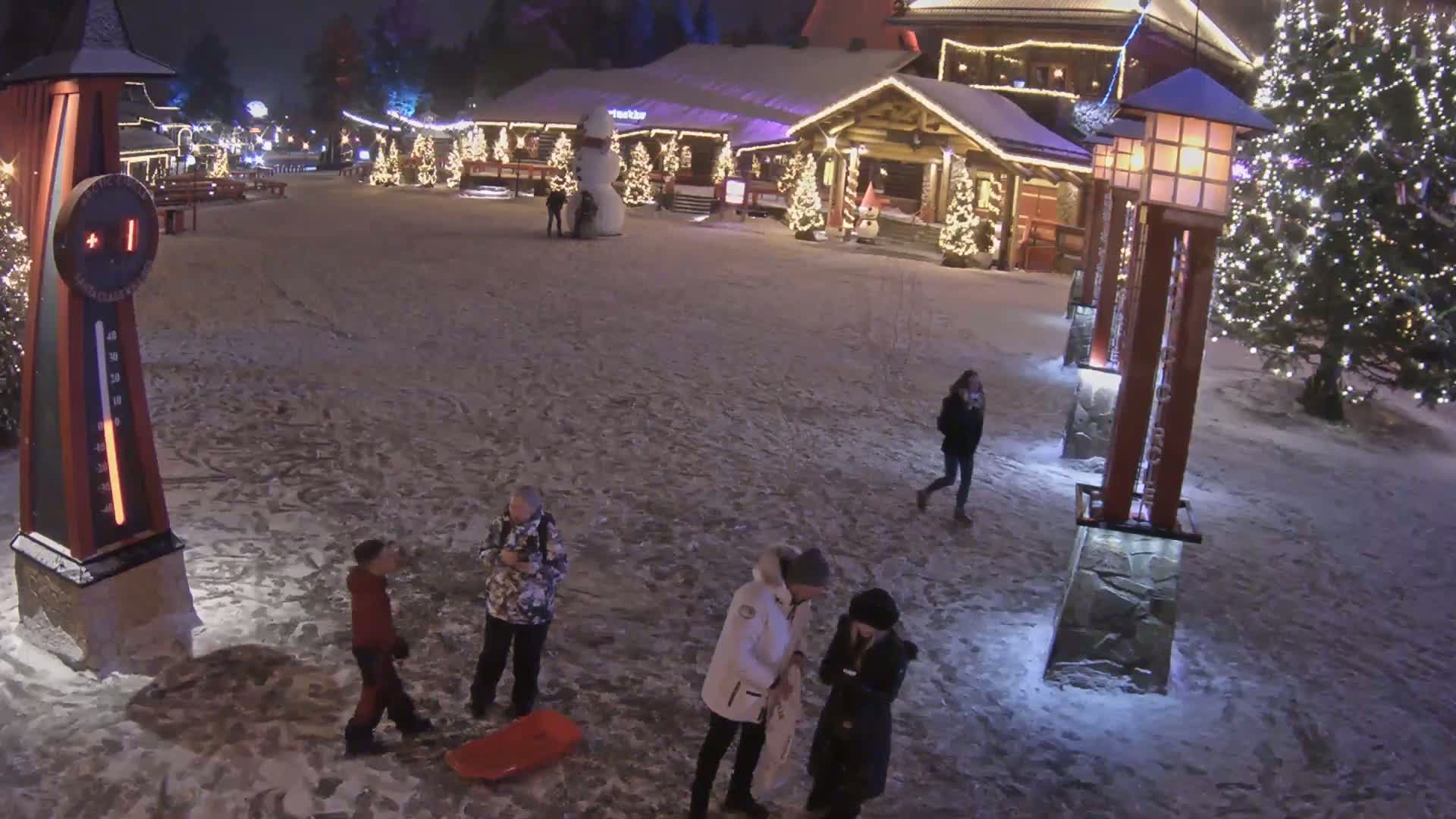 A snow-covered village square at night, illuminated by lights on buildings and lampposts, with several people walking and standing around, one near a red sled.