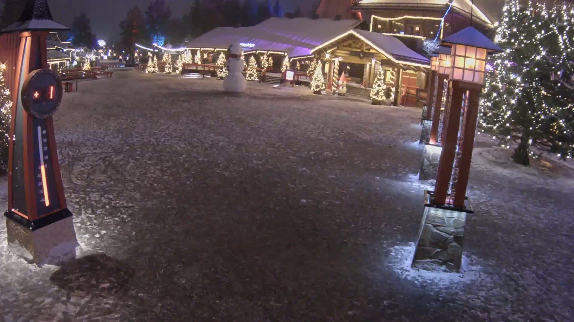 A snow-covered plaza at night features a log cabin-style building decorated with Christmas lights, a snowman, and several illuminated light posts.