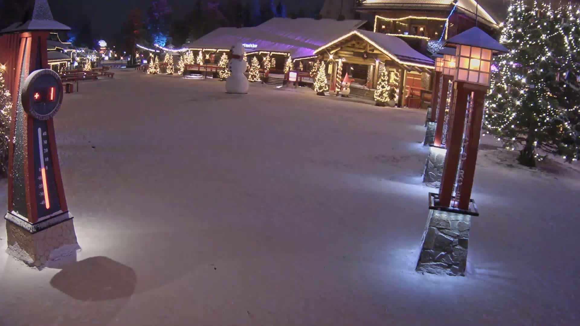 A snow-covered village scene at night, illuminated by lights on buildings and lampposts, features a snowman and a large outdoor thermometer.