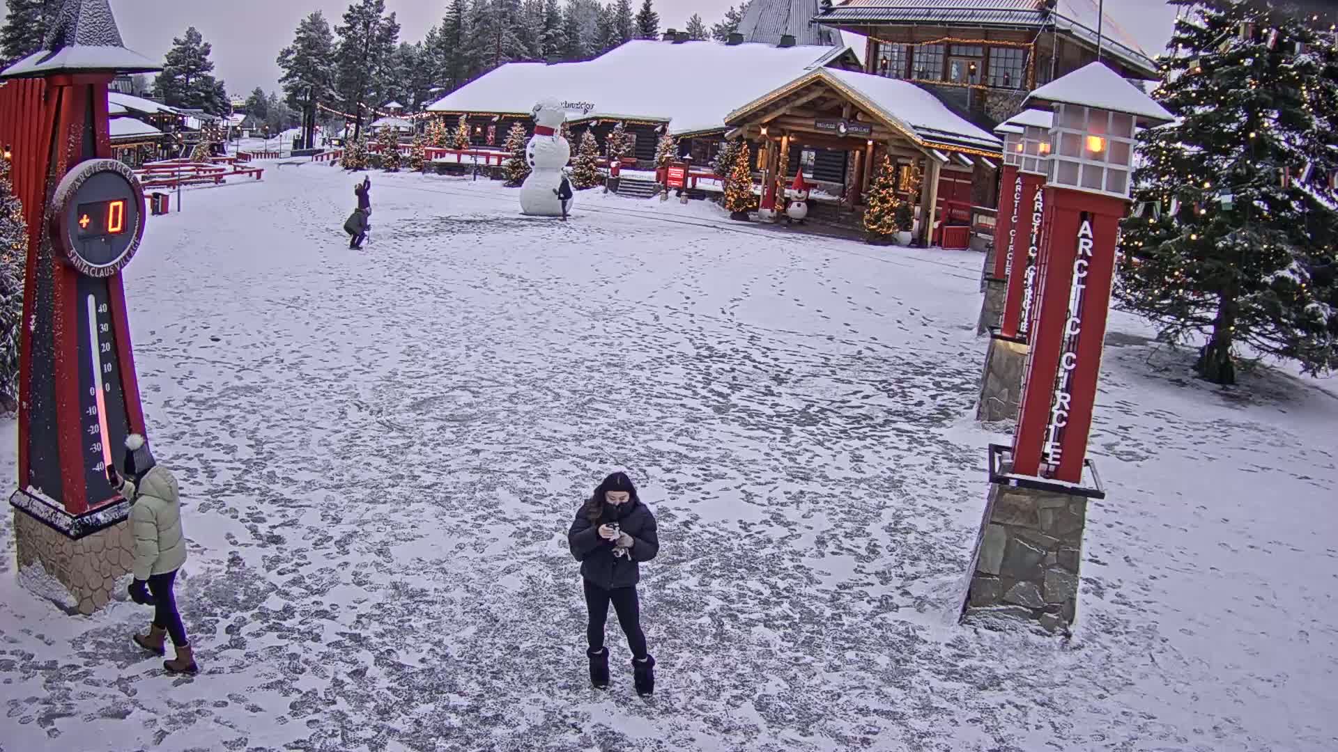 A snow-covered plaza with several people, a large snowman, and festive buildings under an overcast sky.