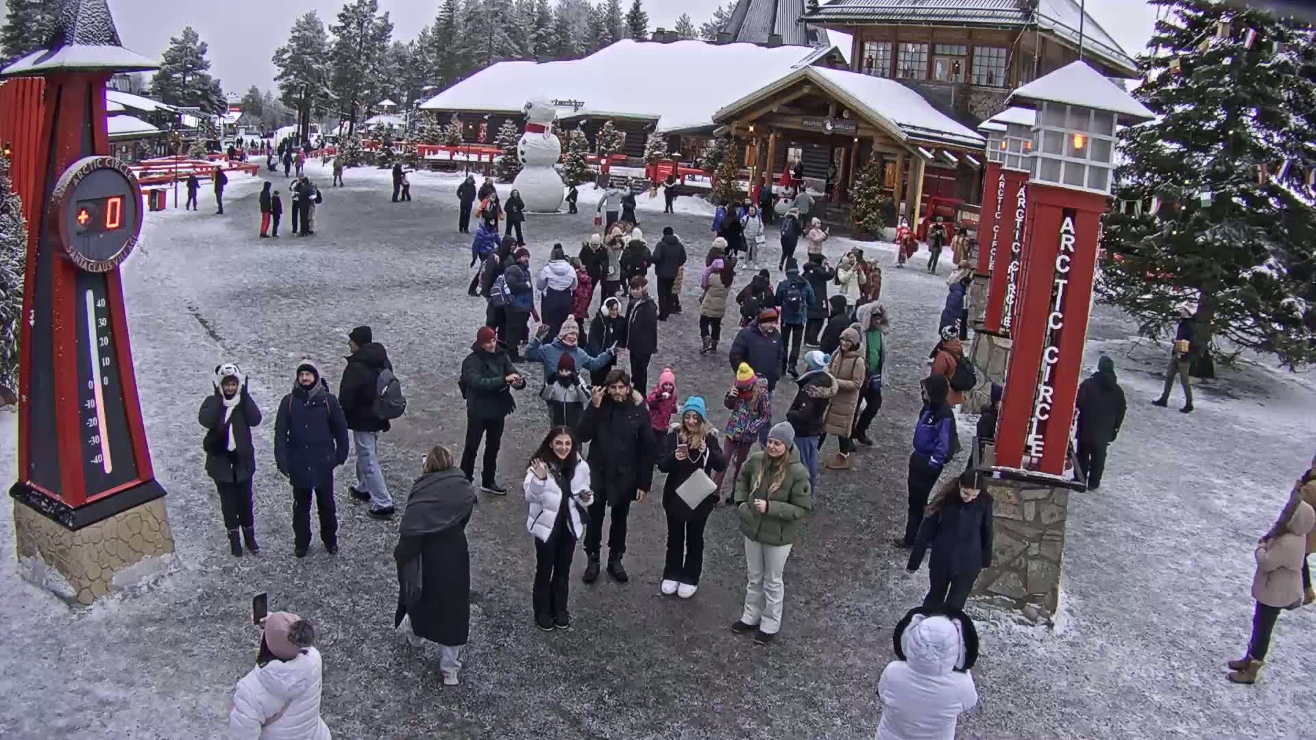 A large group of people gathers in a snowy plaza in front of a building, with a large snowman visible in the background and a thermometer showing a temperature of 0 degrees.
