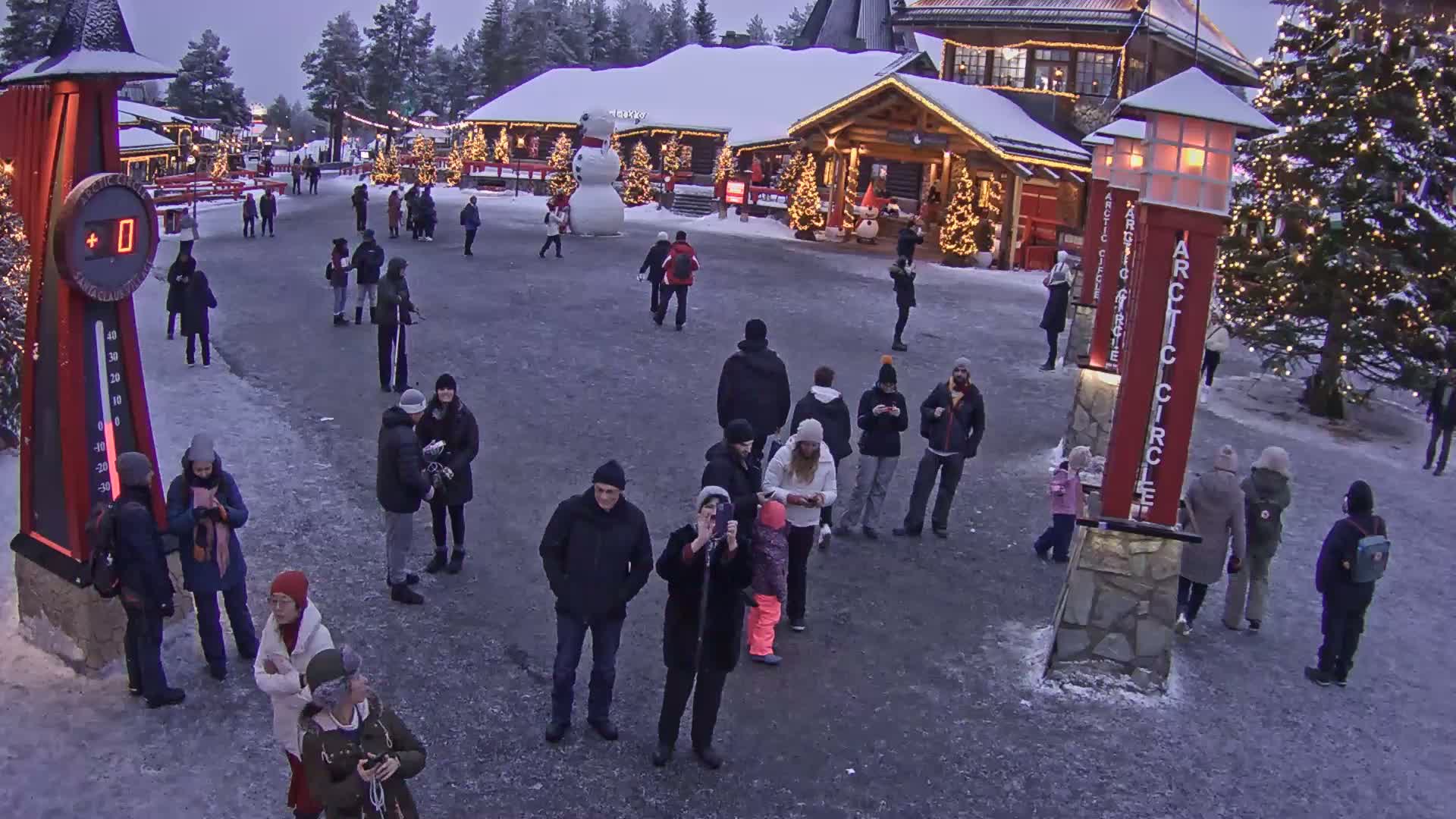 A snowy town square with many people walking around, some standing in small groups, under a twilight sky.