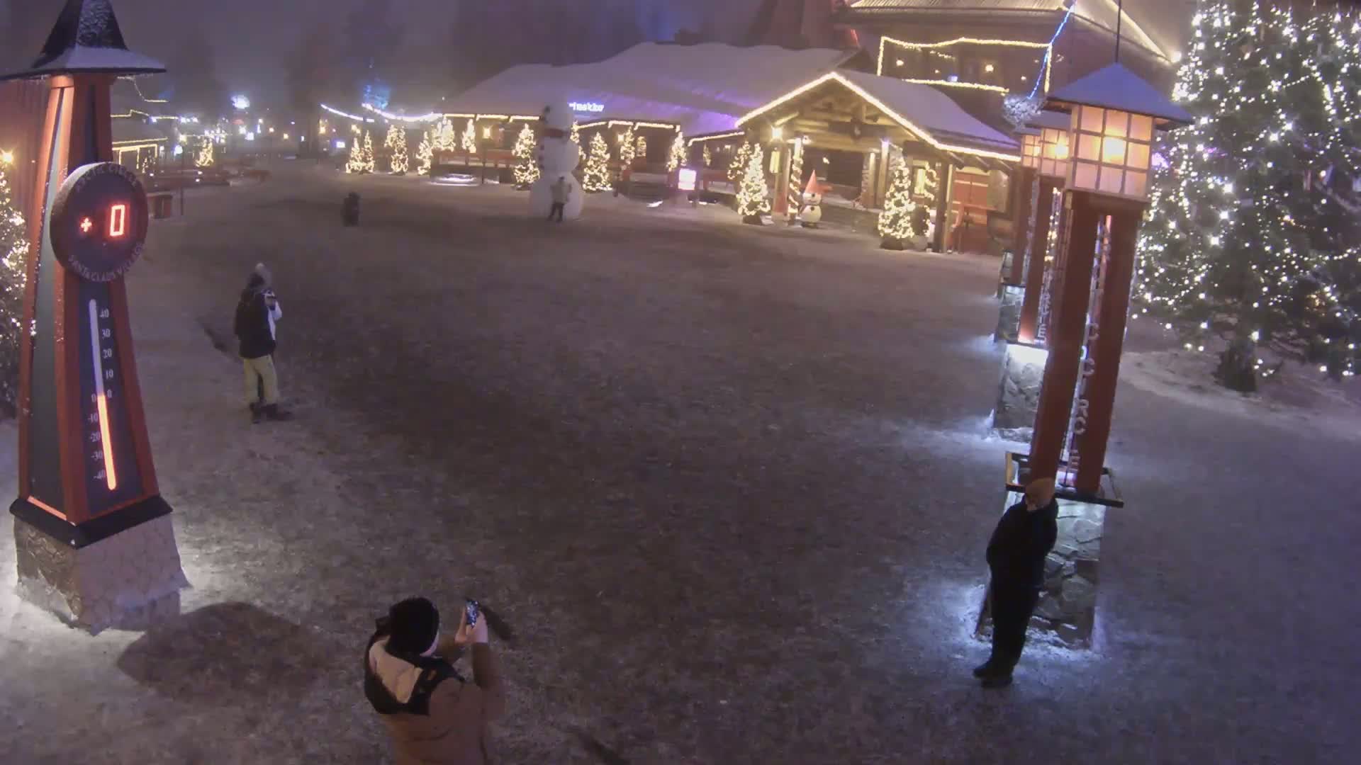 A snowy, nighttime scene shows a plaza with several people, a snowman, decorated Christmas trees, and illuminated buildings.