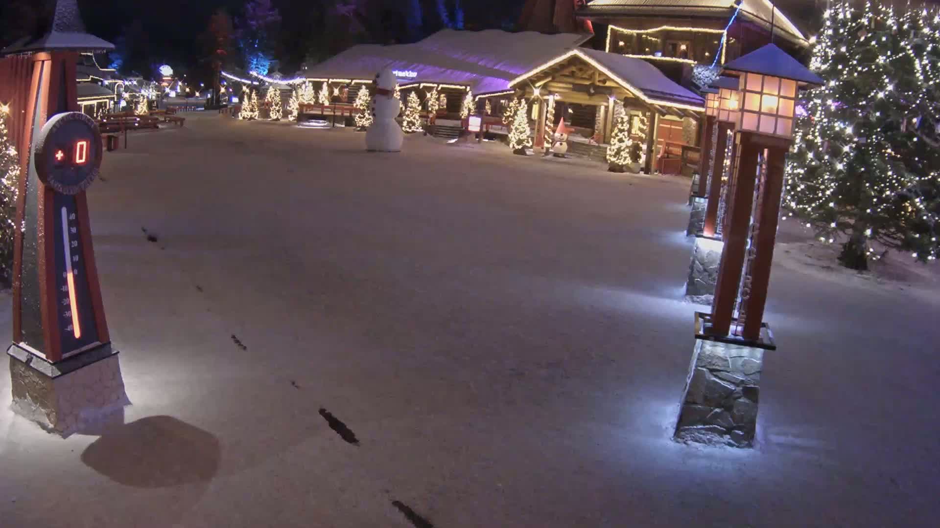 A snow-covered plaza at night features a few illuminated Christmas trees, a snowman, and a lighted building with a thermometer showing 0 degrees.