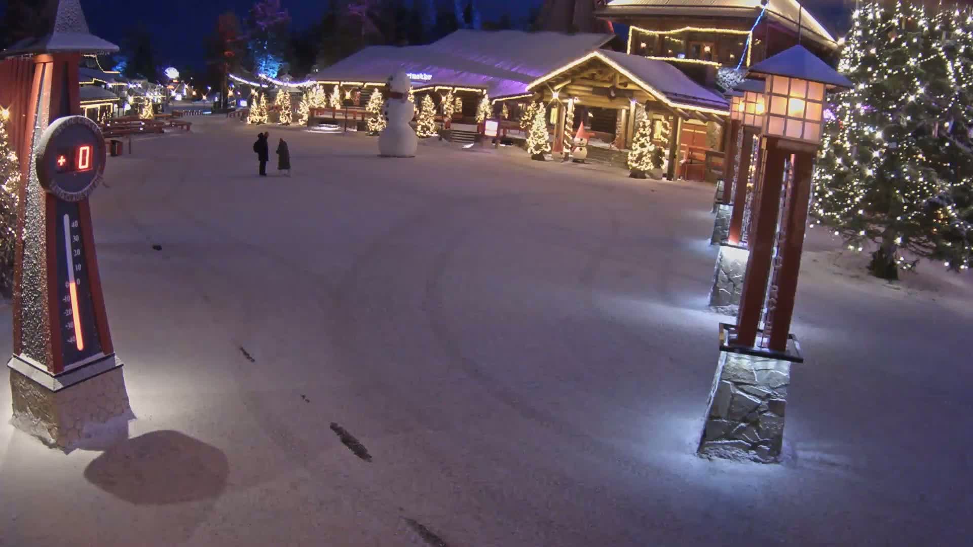 A snow-covered plaza at night, illuminated by lights on buildings and lampposts, features a snowman and a few people walking past a Christmas-decorated building.