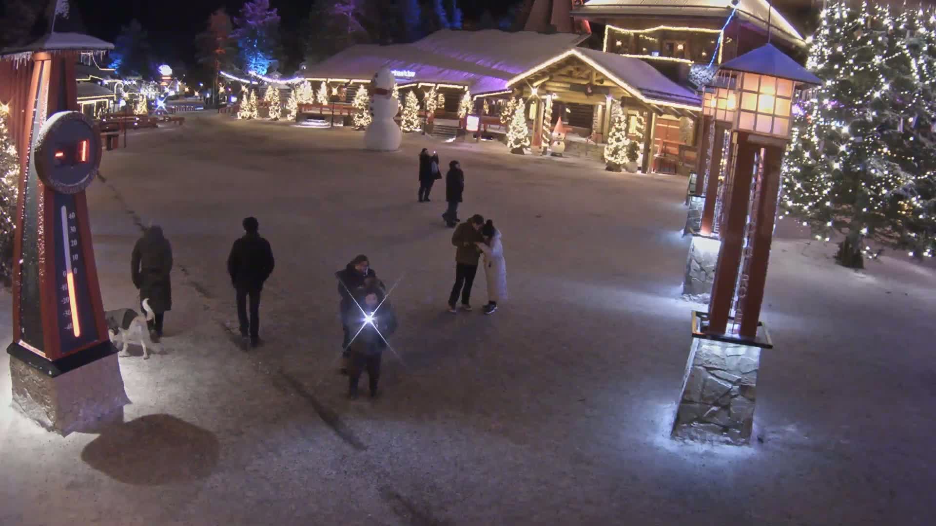 A snow-covered plaza at night features several people walking and standing around, illuminated by decorative lights on buildings and lampposts, with a large snowman nearby.