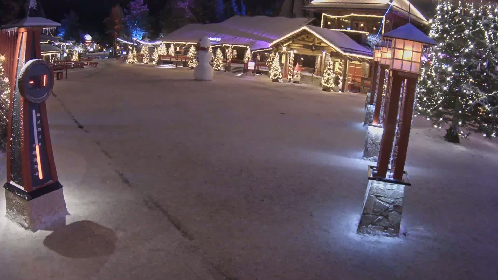 A snow-covered village square at night, illuminated by lights on buildings and lampposts, features a snowman and a large thermometer showing a sub-zero temperature.