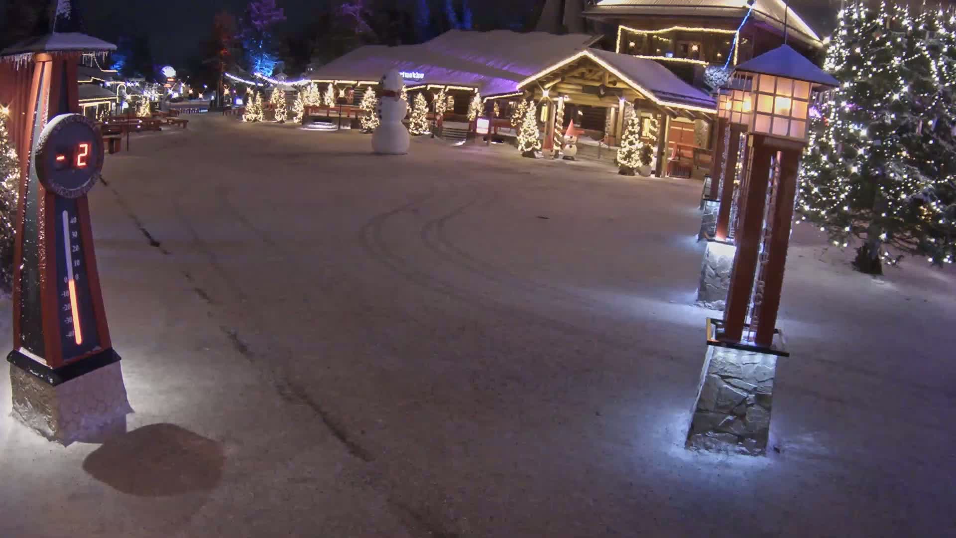A snow-covered nighttime village scene features a snowman, illuminated Christmas trees, and decorative lights, with a temperature of -2 degrees displayed on a nearby thermometer.