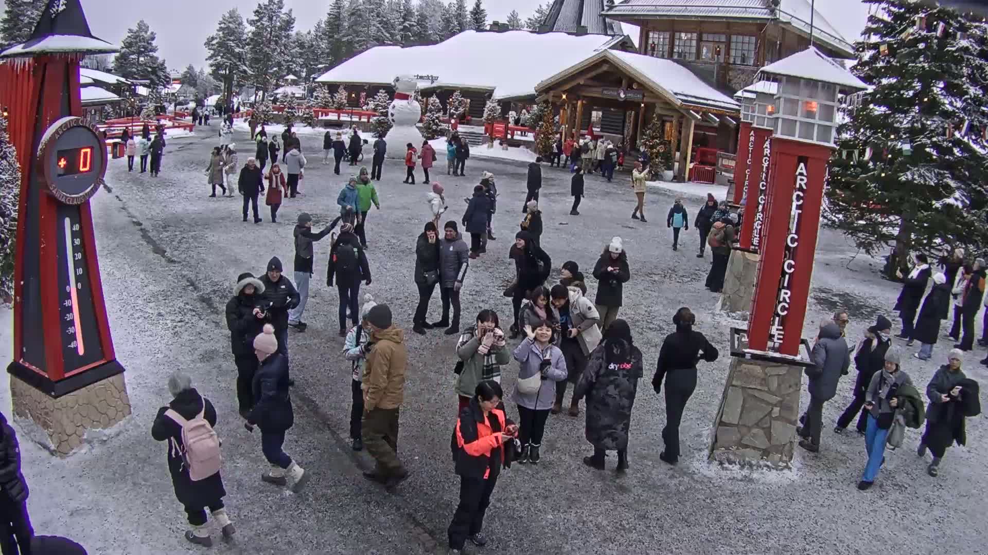 A crowd of people gathers in a snowy plaza in front of wooden buildings, with a large snowman visible nearby.