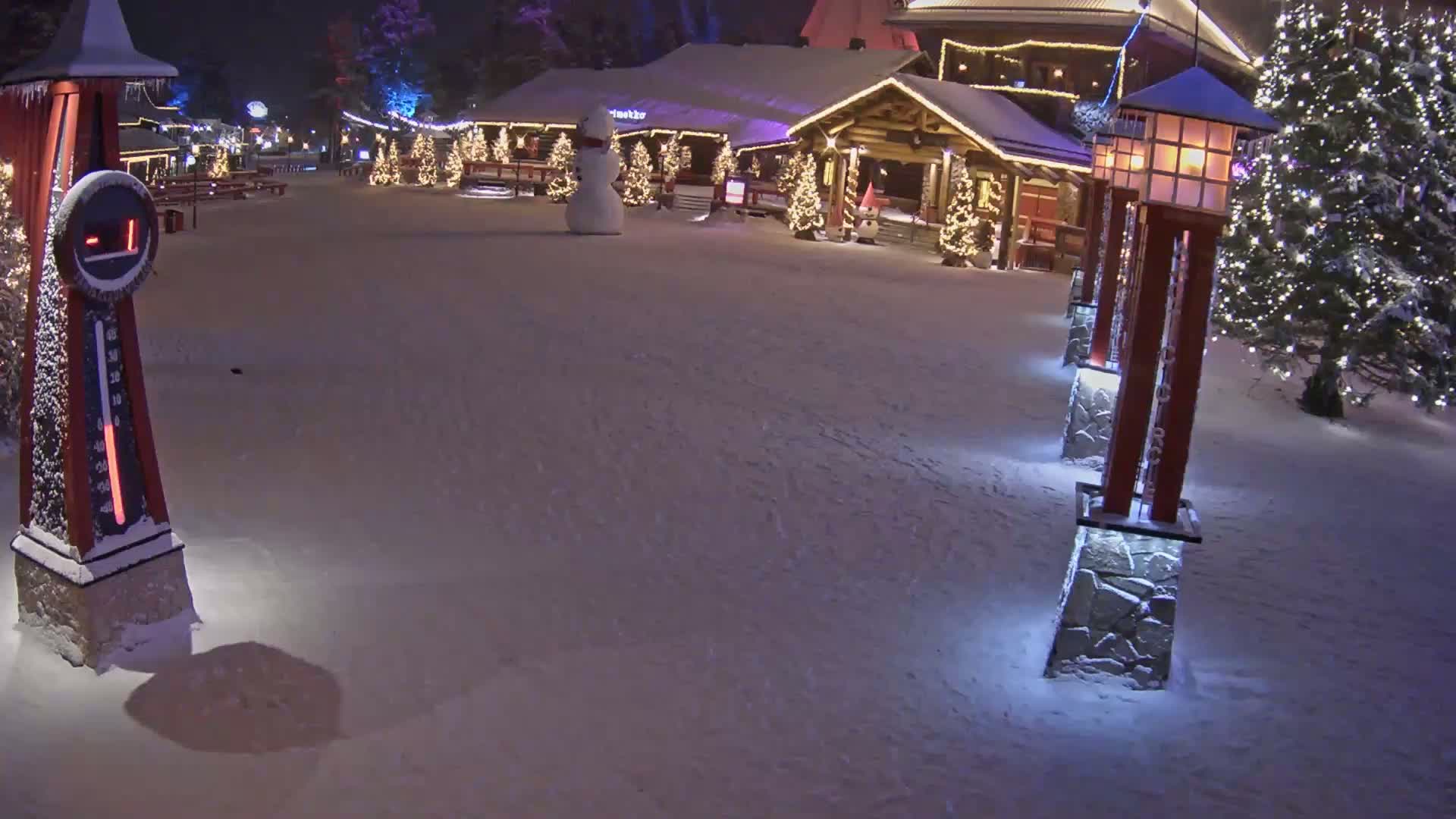 A snow-covered plaza at night shows a snowman, illuminated Christmas trees, and a row of lit lanterns near a wooden building; the temperature is -1 degree.
