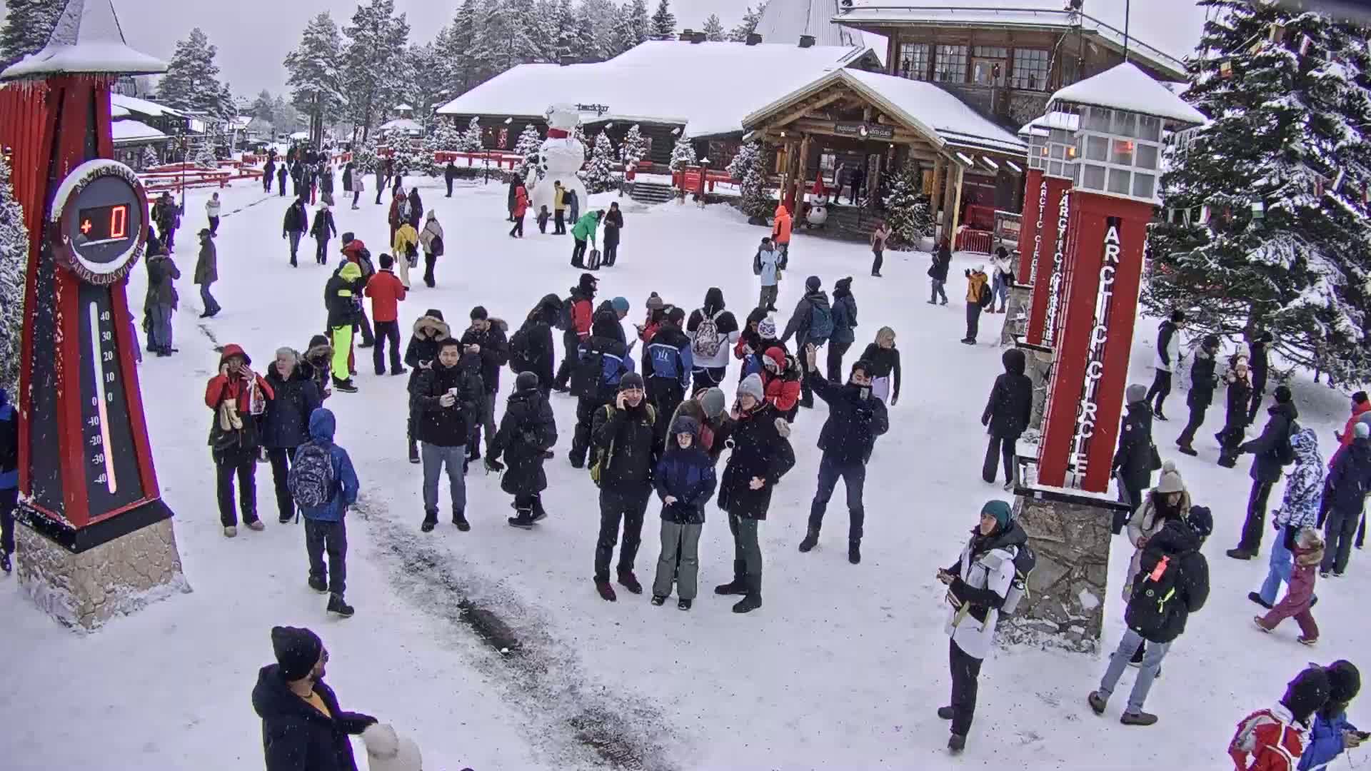 A large group of people are gathered in a snowy plaza in front of a building, with a large snowman visible in the background.