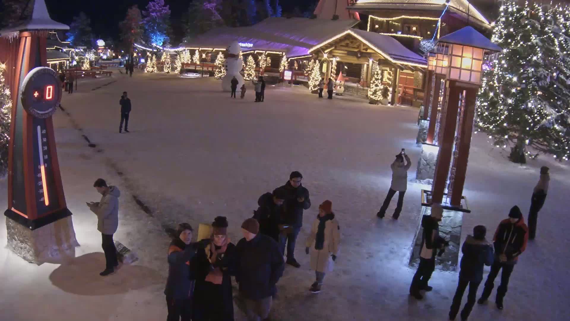 A snow-covered plaza at night, illuminated by lights on buildings and lampposts, features several people gathered around and near a large thermometer displaying 0 degrees.