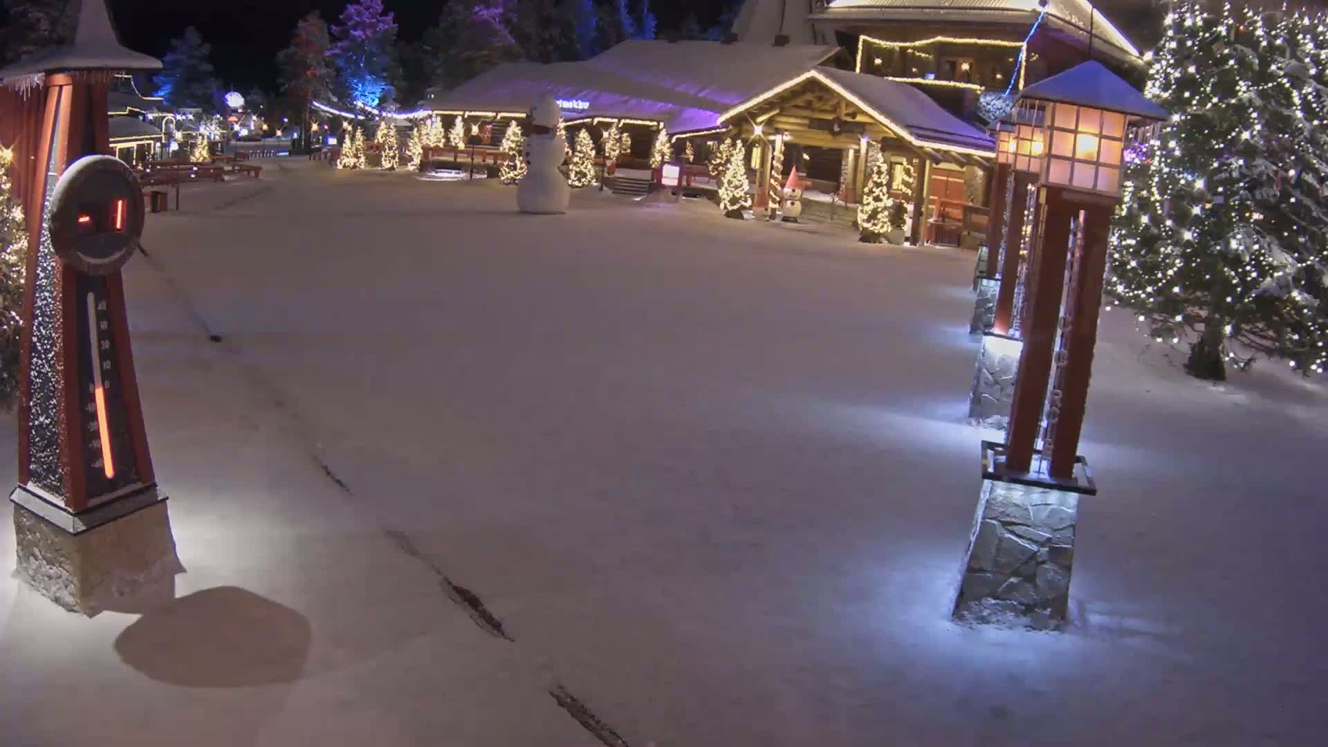 A snow-covered village square at night, illuminated by lights on buildings and lampposts, features a snowman and a large outdoor thermometer.