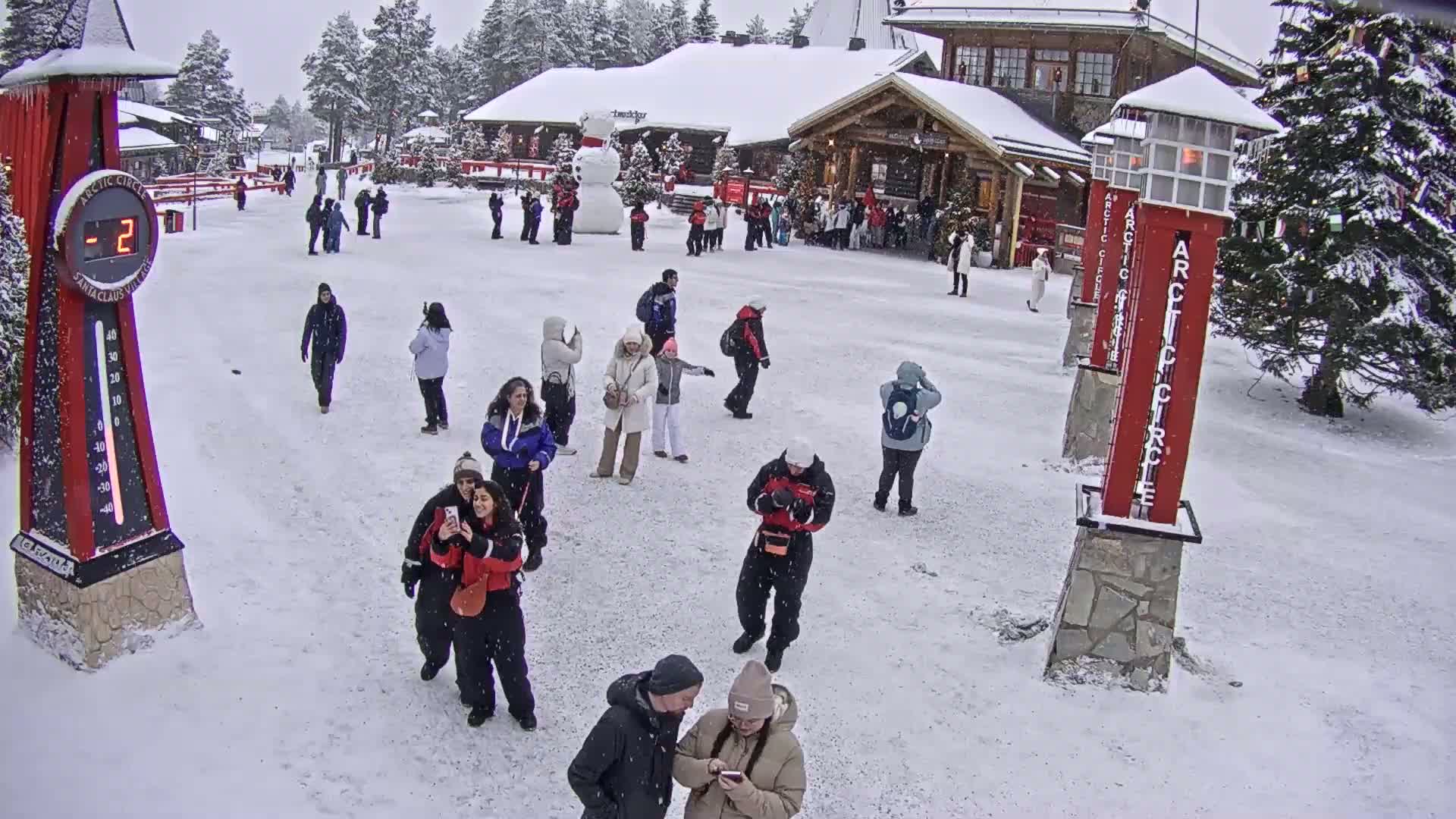 A snow-covered plaza with numerous people walking around near a building, a large snowman, and tall, red Arctic Circle markers; it is snowing.
