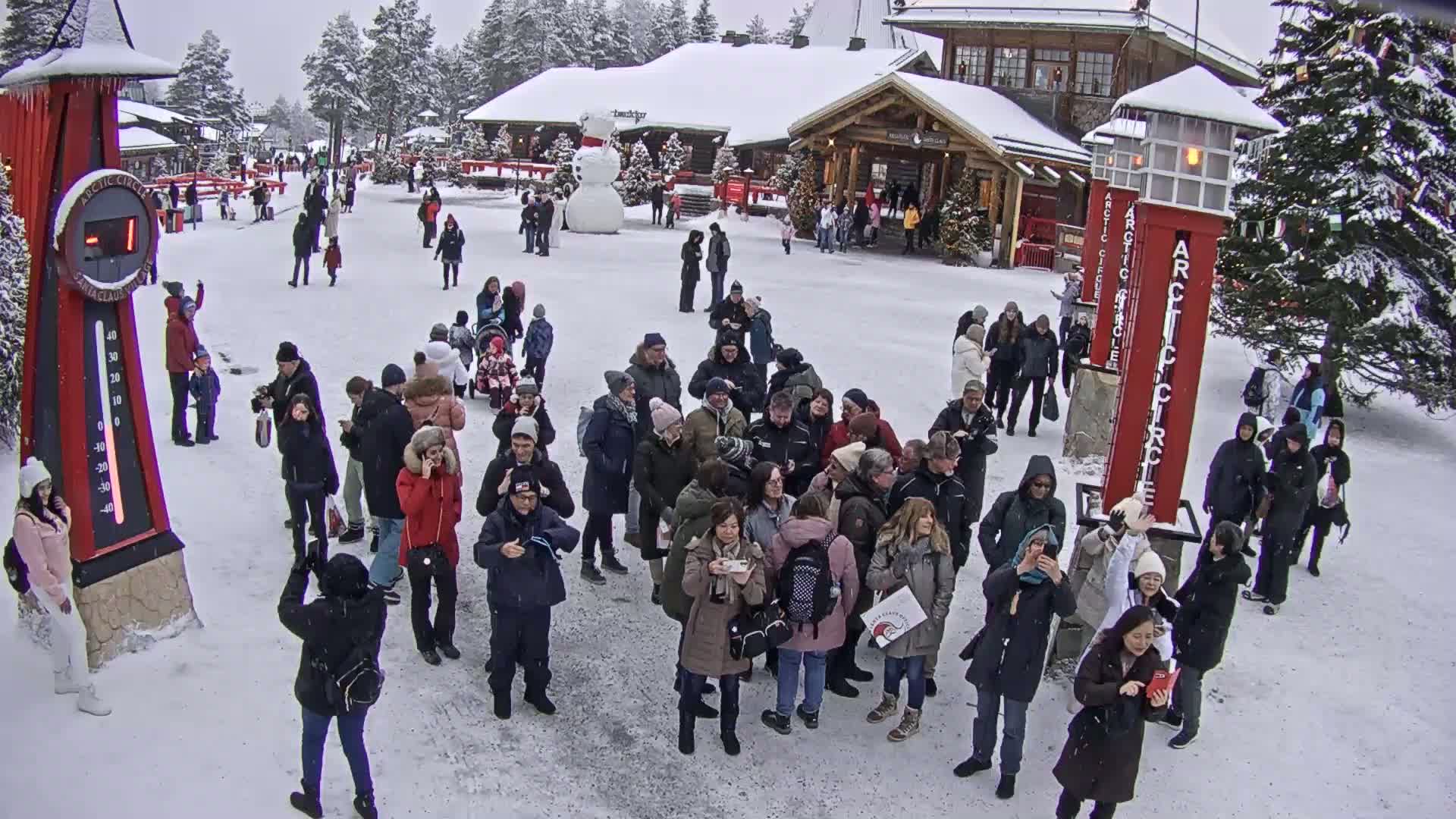 A snowy outdoor scene shows a large group of people gathered in a plaza near buildings, with a snowman visible in the background.