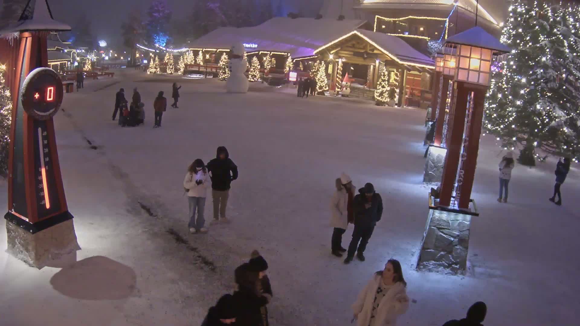A snow-covered plaza at night, illuminated by lights on buildings and trees, with several people walking around in winter clothing near a large thermometer showing a temperature of 0 degrees.