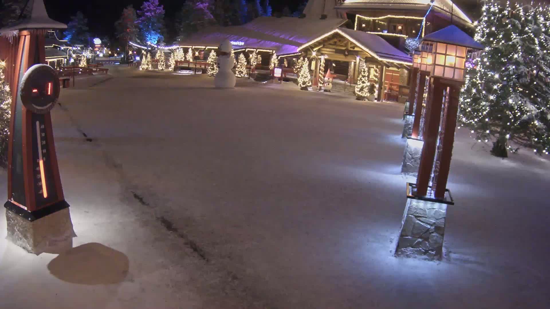 A snow-covered village square at night features illuminated Christmas trees, lit lampposts, a snowman, and a large outdoor thermometer.