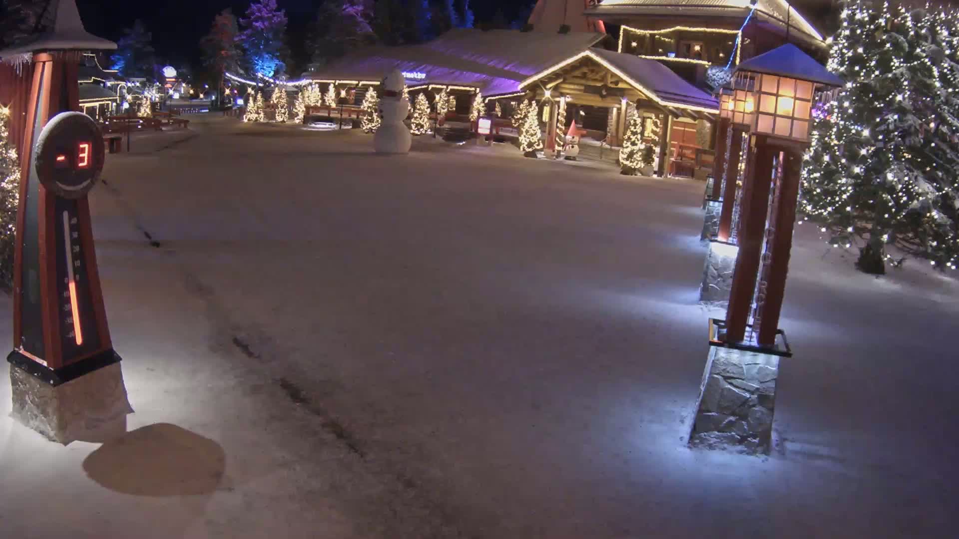 A snow-covered plaza at night, illuminated by lampposts and Christmas lights, features a wooden building and a snowman.