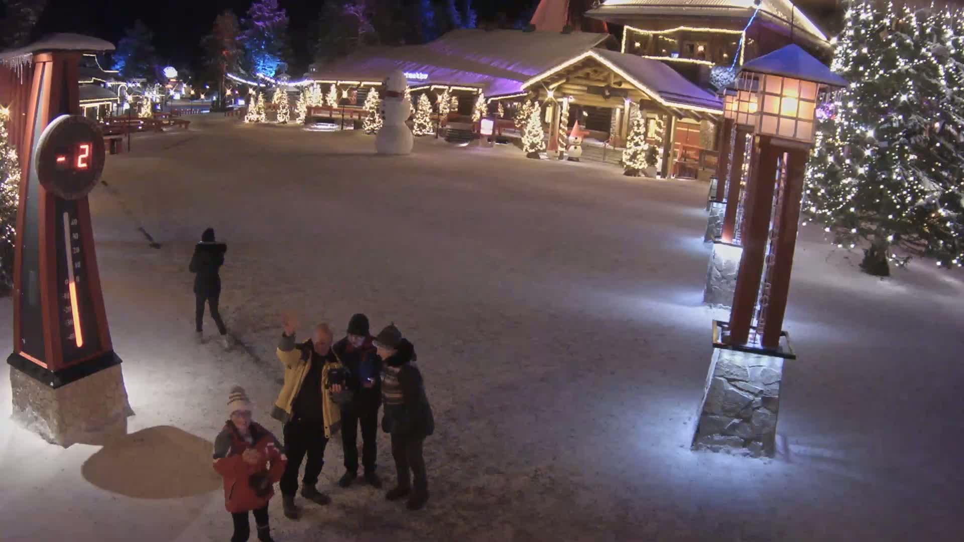 A snow-covered village square at night, illuminated by lights on buildings and lampposts, features several people standing together and a snowman.