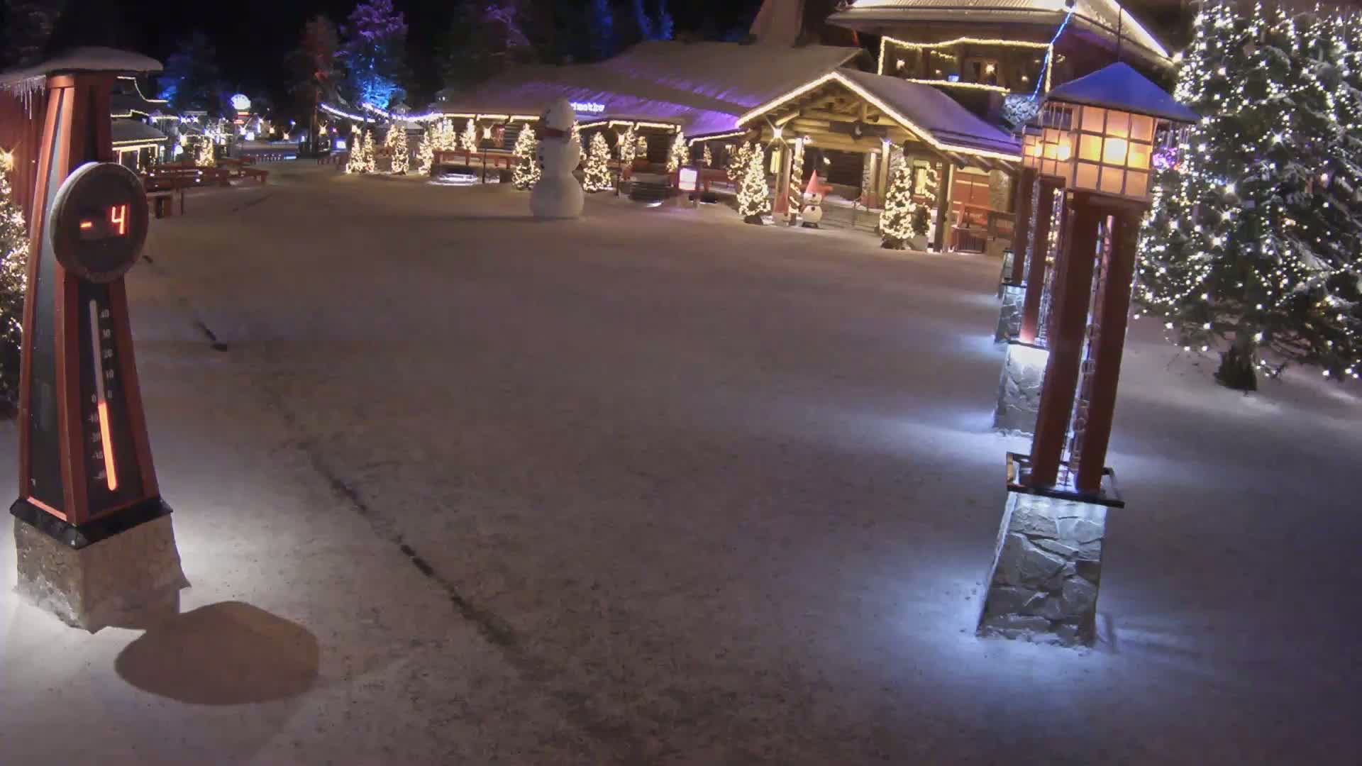 A snow-covered village scene at night, illuminated by lights on buildings and lampposts, features a snowman and shows a temperature of -4 degrees.