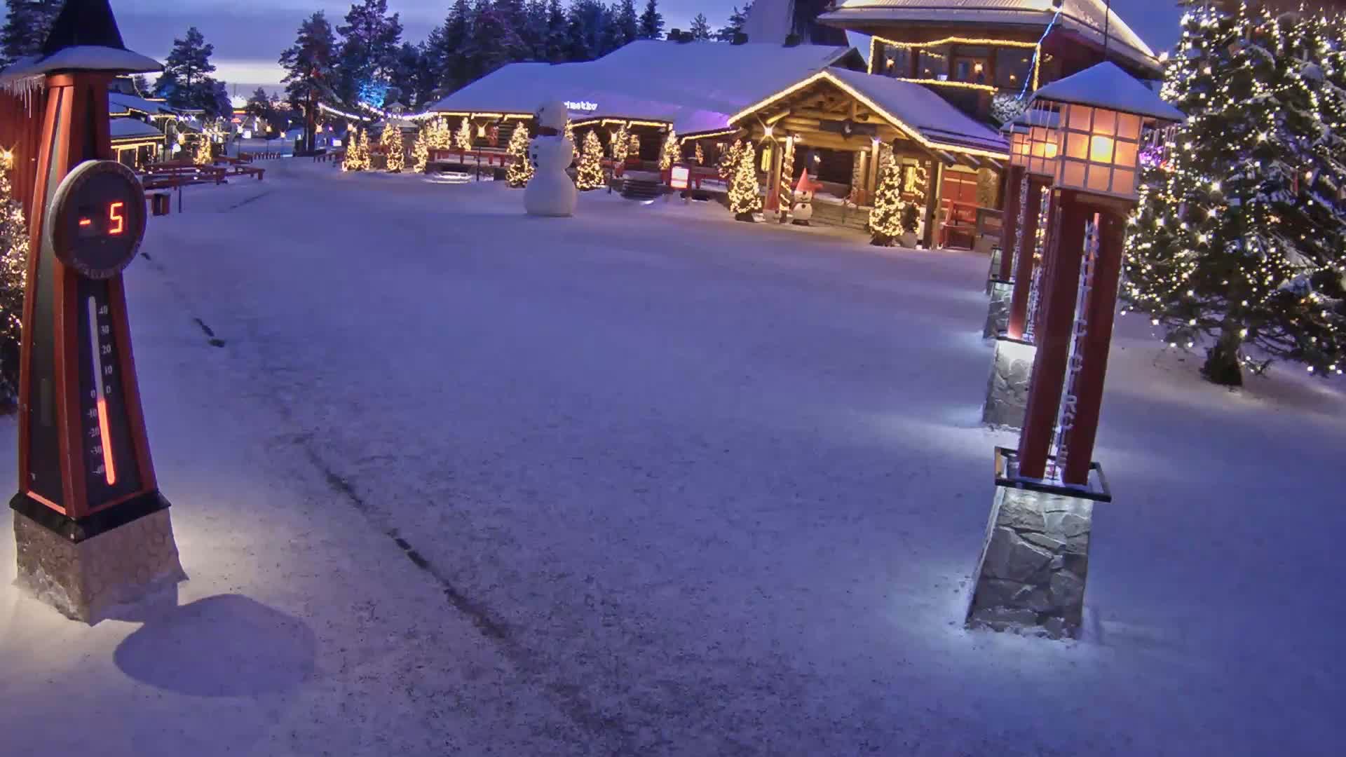 A snow-covered village square at night, illuminated by lampposts and Christmas lights, shows a snowman near a building and a thermometer displaying a temperature of -5 degrees.