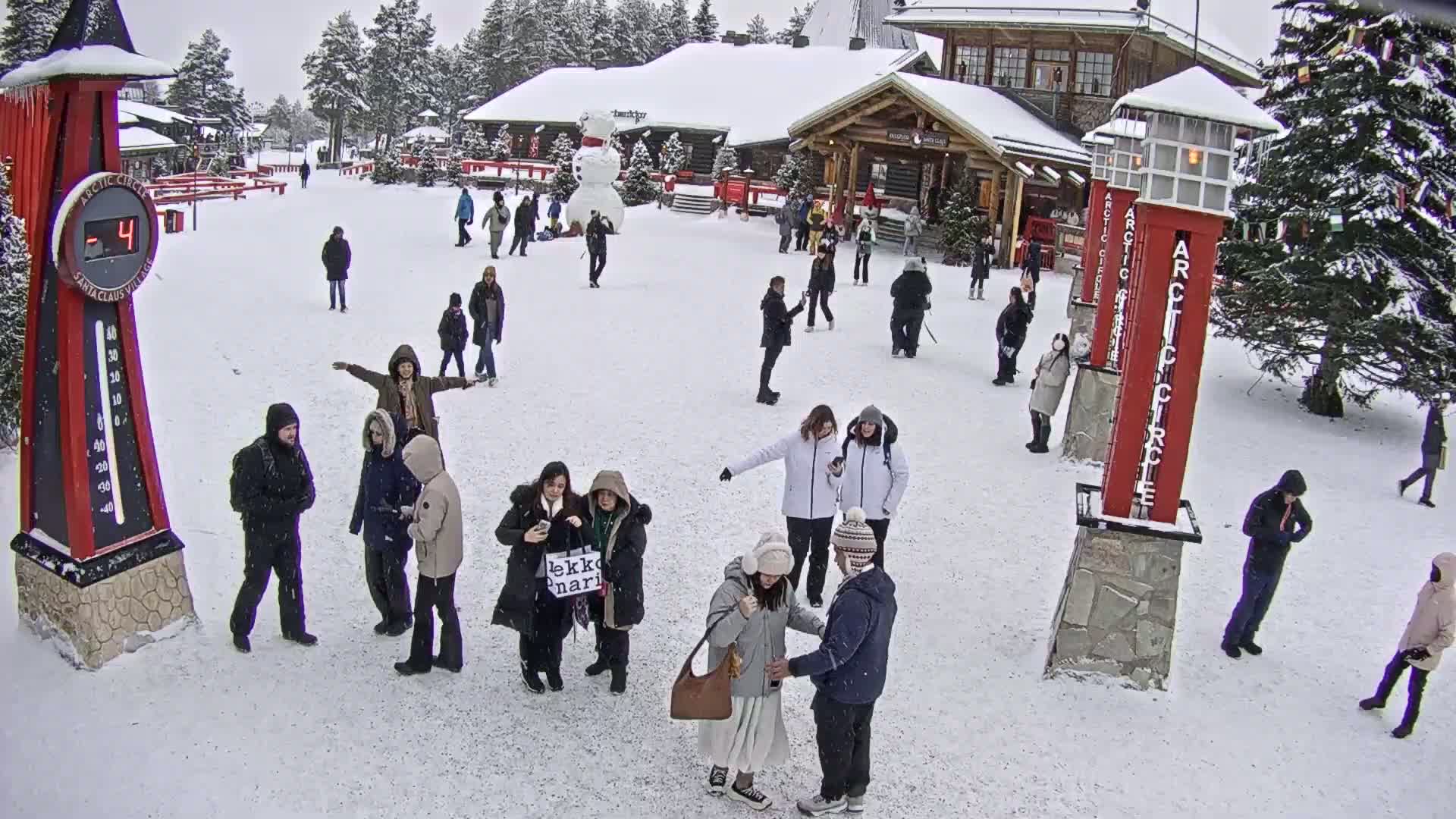 A snowy outdoor scene shows numerous people gathered in a village square, near buildings, a large snowman, and tall red Arctic Circle markers, with a temperature display showing -4 degrees.